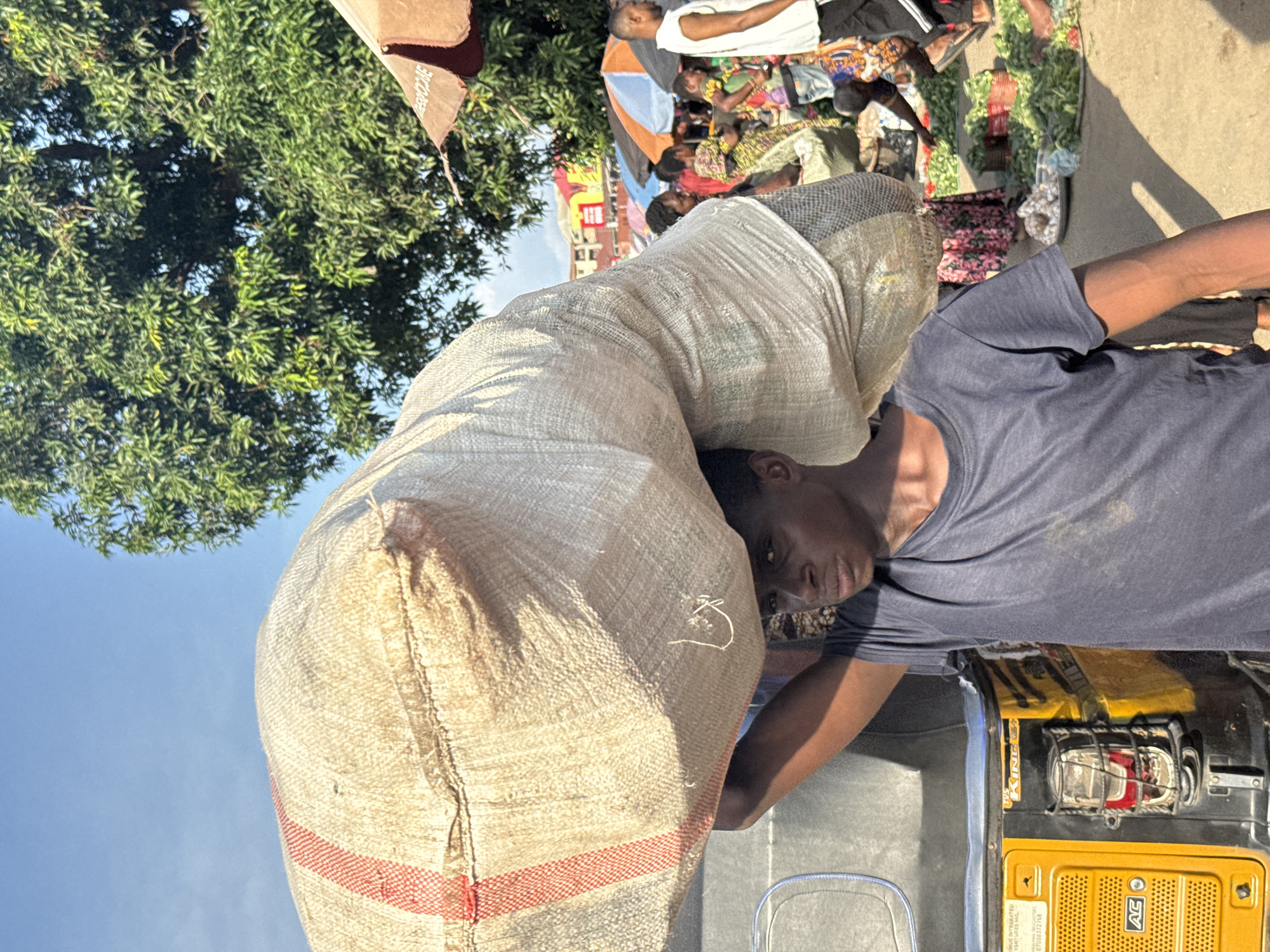 A young man is carrying a large sack of goods on his head in the Ochanja Market in Onitsha, Nigeria. The sack is made of rough, woven material and has two red stripes running vertically along its length. He is wearing a dark blue t-shirt. To his left is the side of a yellow and black auto rickshaw, commonly known as an "okada" or "keke napep," which is often used for transportation and goods delivery in Nigeria. The market appears busy, with other people visible in the background, some of whom are wearing colorful clothing. Several market stalls and umbrellas can be seen, along with piles of green vegetables and other goods laid out for sale on the ground. The sky is clear and blue, indicating a sunny day. The overall scene depicts a typical day in a bustling Nigerian market.