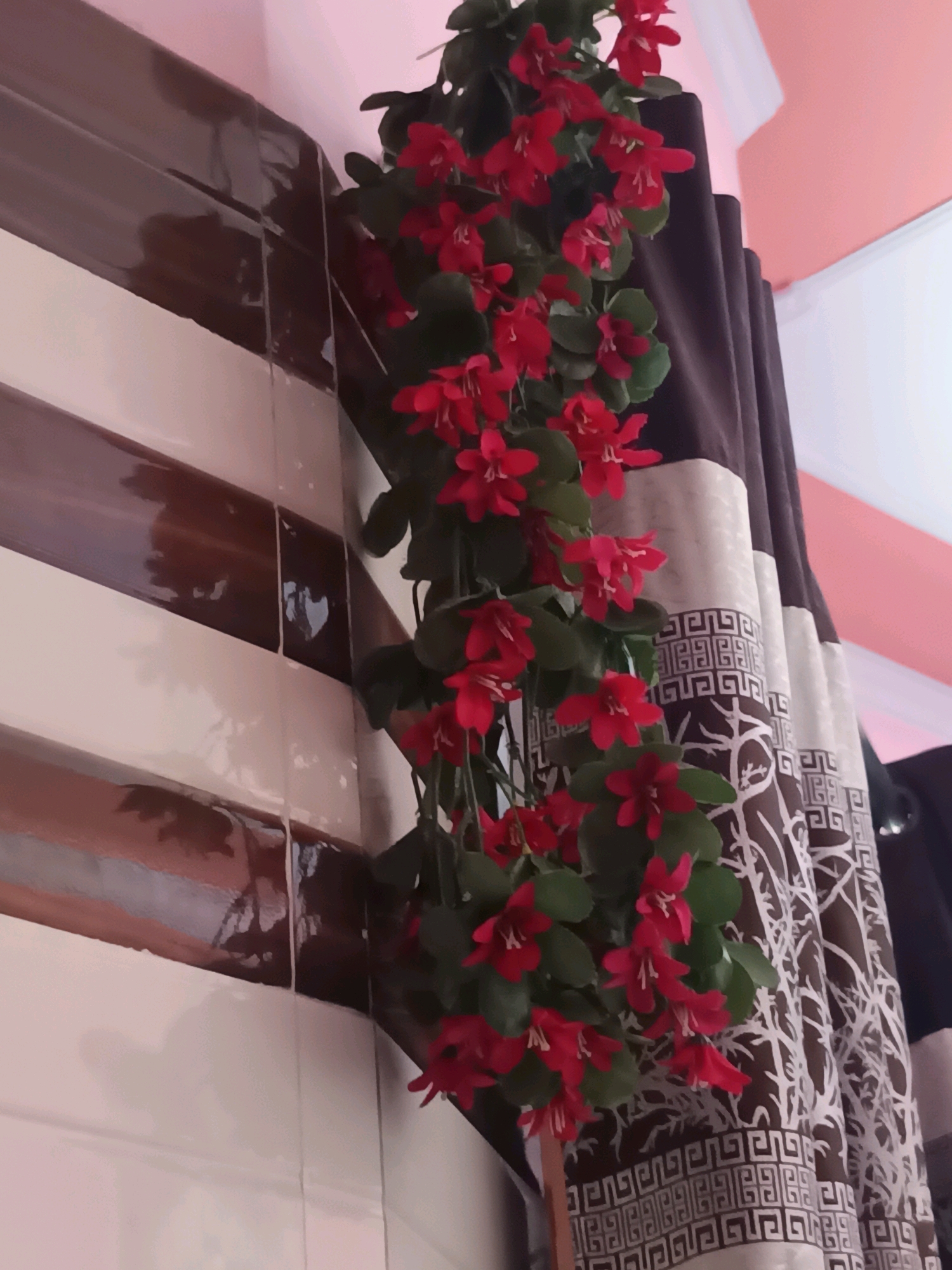 An artificial vine with red flowers and green leaves hangs down a tiled wall. The tiles are a mix of white and dark brown stripes. To the right of the vine, a dark brown curtain with a white pattern hangs. The background appears to be a light pink wall. There are no people or actions depicted. The location is a room in Vijaypur, India.