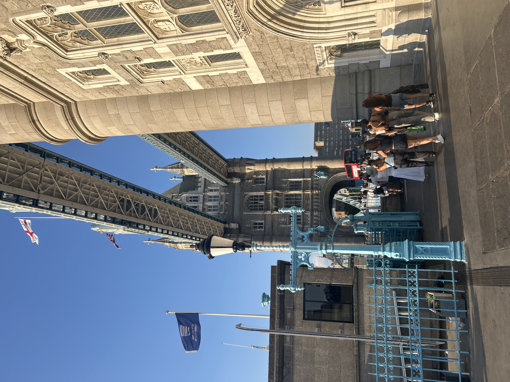 The image shows Tower Bridge in London, United Kingdom, on a clear day. The camera angle is from the walkway of the bridge looking towards the north towers. Two Union Jack flags are visible flying from the bridge structure, as well as a blue flag with white text that reads "CITY BRIDGE FOUNDATION". A group of people are walking away from the camera on the bridge's pedestrian walkway. They are casually dressed and appear to be tourists. In the background, a red double-decker bus is visible, along with other buildings. The lighting suggests it is late afternoon or early evening, with long shadows cast by the bridge and people. The architecture of Tower Bridge, with its stone towers and intricate gothic-style windows, is a prominent feature of the scene. The blue decorative fence and lamppost on the left side of the image add to the distinct visual elements of the London landmark.