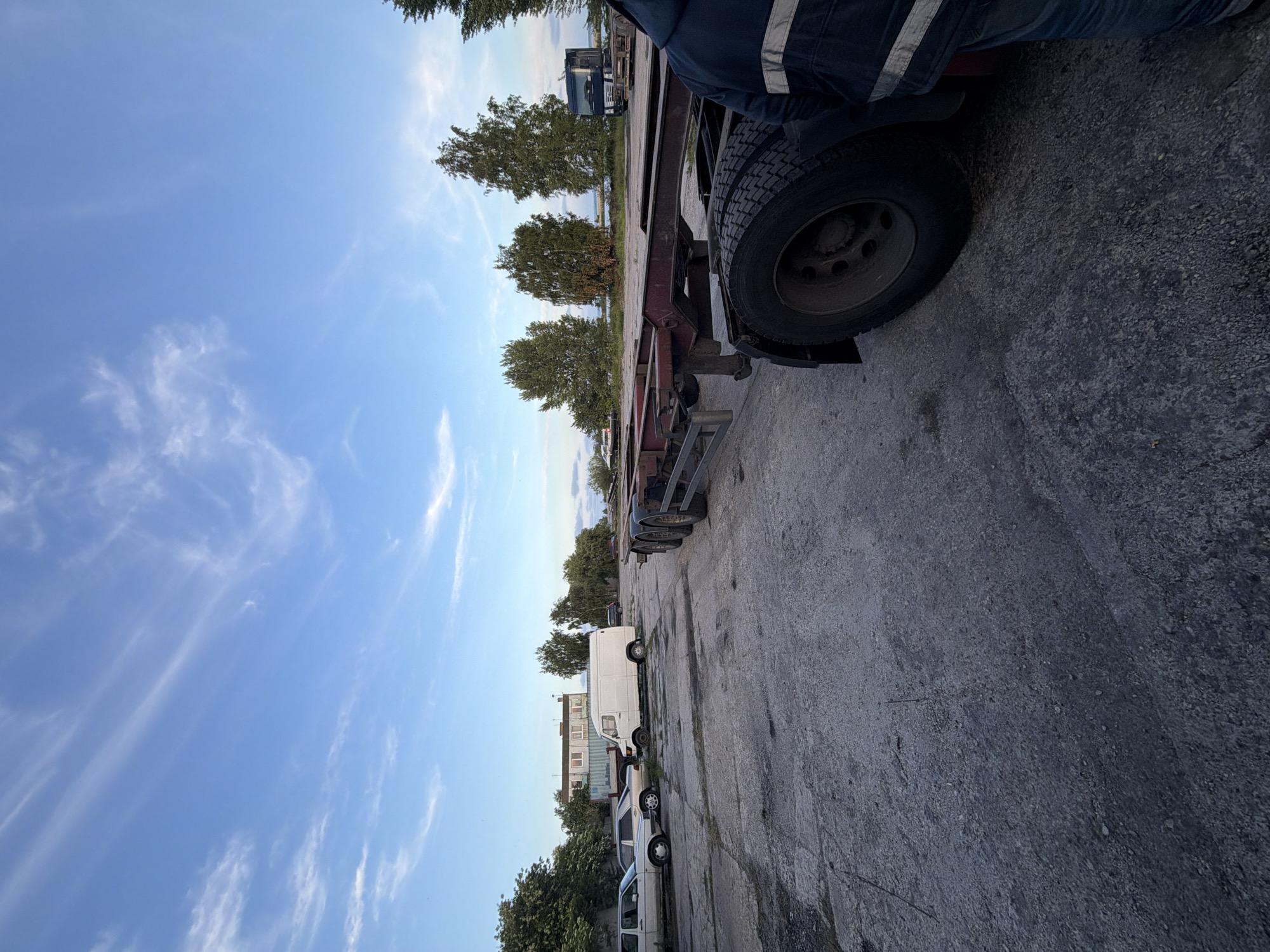 The image shows a low-angle view of a flatbed trailer parked on a gravel lot, with a clear blue sky and sparse white clouds above. A person in work clothes, with reflective stripes on their jacket, stands next to the trailer's tire. The trailer is dark red and appears to be empty. In the background, several vehicles are parked in a line, including a white van and two cars. Trees and buildings can be seen behind the parked vehicles, suggesting an industrial or commercial area. The lighting indicates it is daytime, likely late afternoon or early evening due to the angle of the sun and the slightly muted colors. The scene is static, with no active event taking place. There is no visible text in the image. The location is identified as Baltiysk, Russia, which could imply a port or industrial setting, but this is not directly evident from the image itself. The overall mood is neutral and observational.