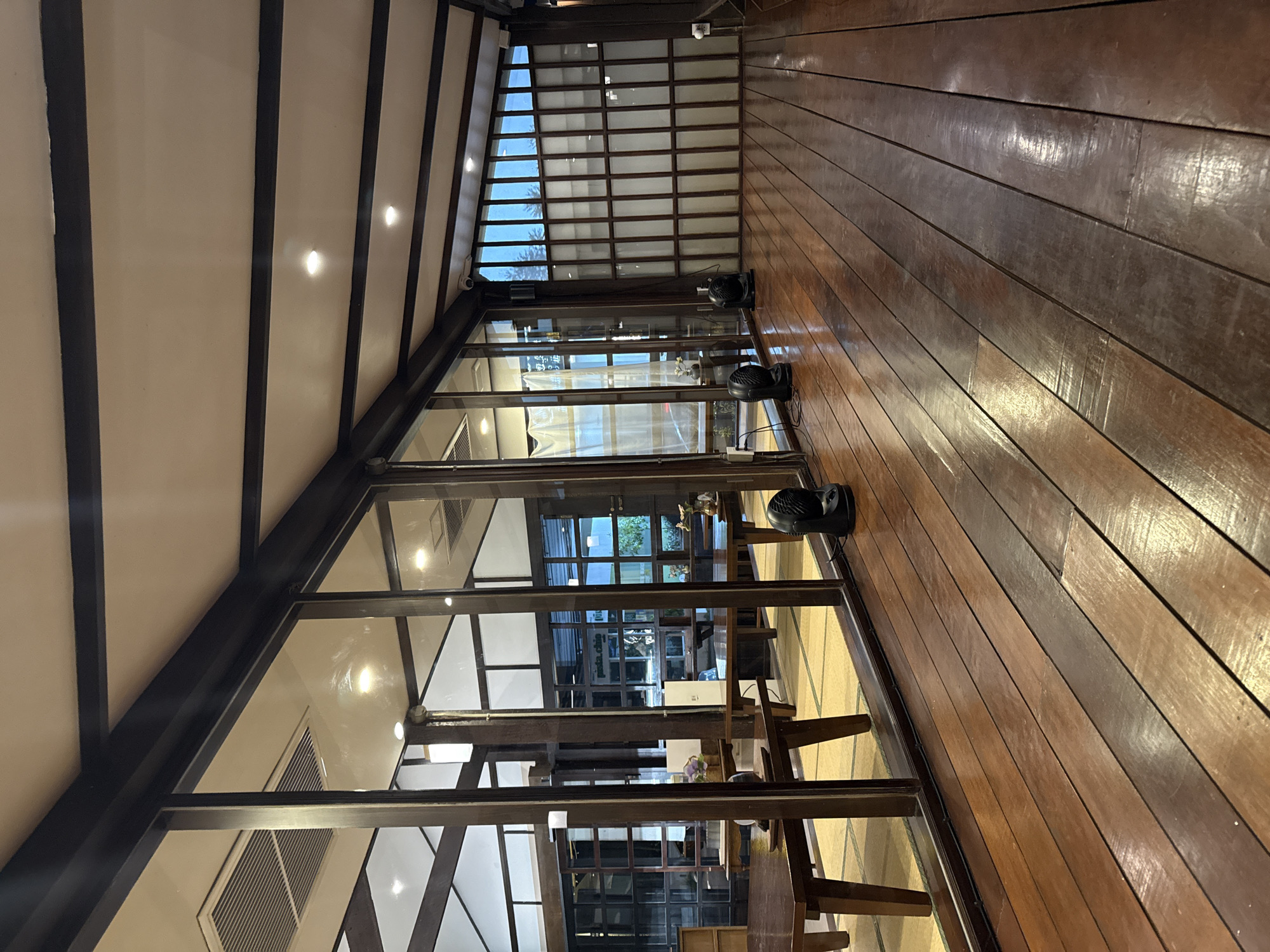 The image depicts an interior space, likely in Chiang Mai City Municipality, Thailand. The structure has a distinctive sloped ceiling with dark beams and a light-colored surface between them. Large windows with dark frames provide views into another room with traditional Japanese seating on tatami mats. Three black fans are positioned along the edge of the wooden floor, which is made of narrow, dark-toned planks running horizontally. The natural light from outside is entering the space through the windows.