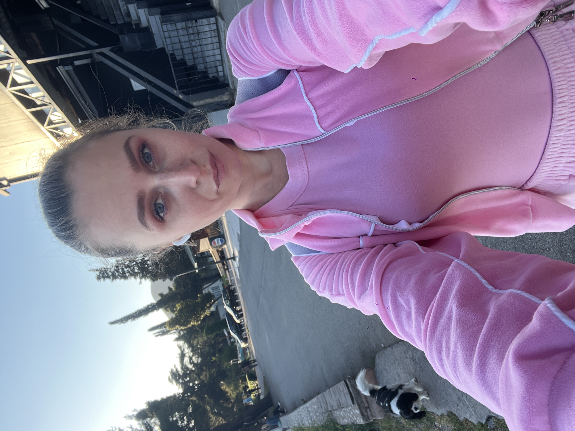 This vertically oriented selfie captures a person in a bright pink velour tracksuit, taken outdoors in Budva, Montenegro, under clear daylight conditions. The image is rotated, presenting a tilted perspective.
The primary subject is a woman with light-colored hair pulled back, blue eyes, and a nose piercing. She wears a full two-piece tracksuit, consisting of a zip-up jacket over a matching pink top and elasticated-waist pants, all made of a velour-like fabric. The tracksuit features white or light grey piping details along the seams and edges. Her expression is neutral as she looks towards the camera.
In the lower foreground (when viewed upright), a small, black and white dog, possibly a Border Collie mix, is lying relaxed on the ground near the person's feet.
The background depicts an outdoor, possibly urban or semi-urban environment. On one side, there are modern buildings with dark roofs, featuring architectural structures like metal trusses and grates. A clear, bright blue sky is prominent above. On the opposite side, a street or paved area is visible with parked cars, trees, and distant buildings. A faint "D" logo can be discerned on a building in the far background. The scene suggests a casual moment during a sunny day.