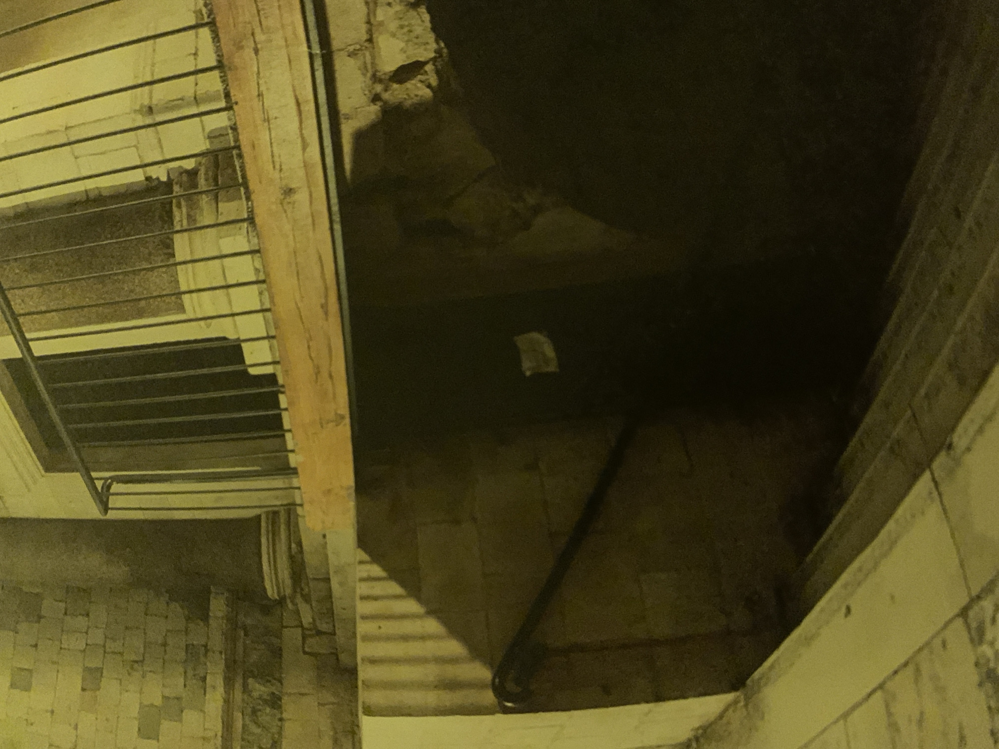 An artificially illuminated, poorly exposed photograph shows a staircase leading into a dark chamber. Visible are the edges of steps and a rough stone floor, with a metal railing at the top of the steps. There are some indications of brickwork along the edges of the scene. The environment is dim and appears to be underground. The location is identified as Barcelona, Spain.
