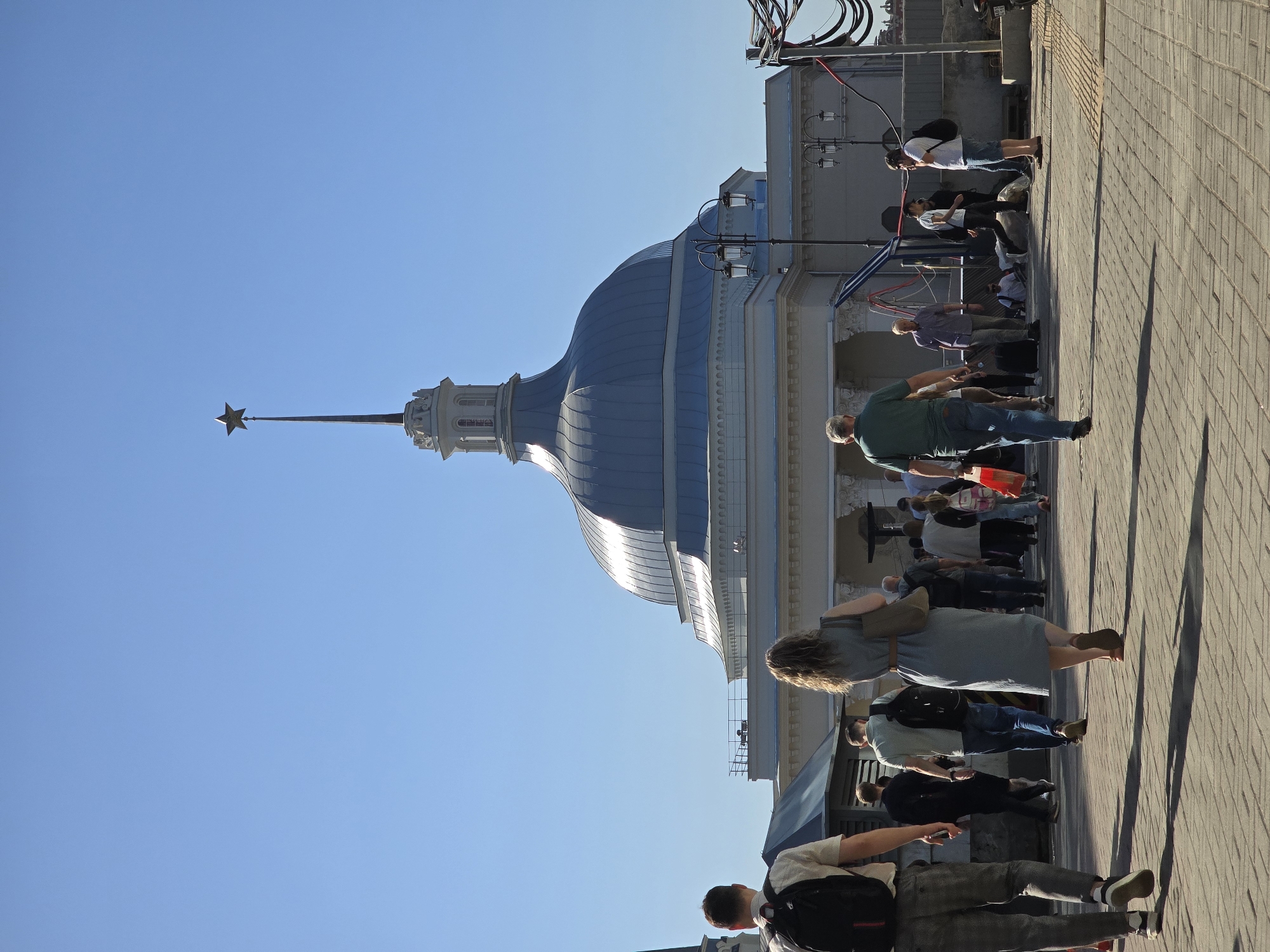 The image captures a scene near the Leningradsky railway station in Moscow, Russia. The most prominent architectural feature is the dome of the station building, topped with a spire and a red star. A group of pedestrians walk along a paved area adjacent to the station. They are mostly casually dressed. Shadows of the people stretch across the paving stones. Utility wires are visible near the building. The sky is clear and blue.