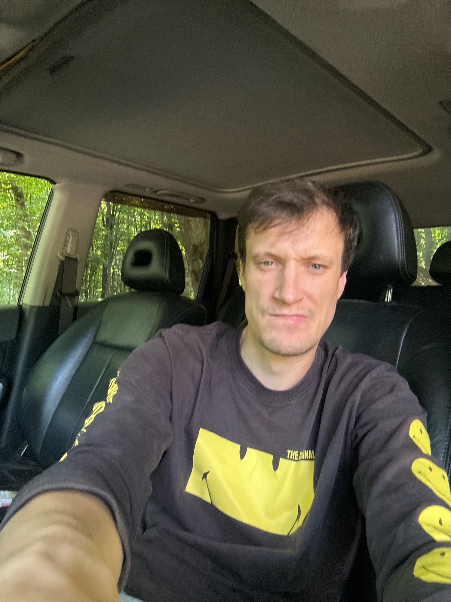 A man with brown hair and a light complexion is sitting in the driver's seat of a car. He is wearing a black long-sleeved shirt with yellow smiley faces printed on it and the text "THE ANIMAL" on the right sleeve. The car has black leather seats and a sunroof. Outside the car, trees and foliage are visible, suggesting a natural, possibly wooded, environment. The image appears to be a selfie taken from the driver's perspective. The location is identified as Shatava, Ukraine.