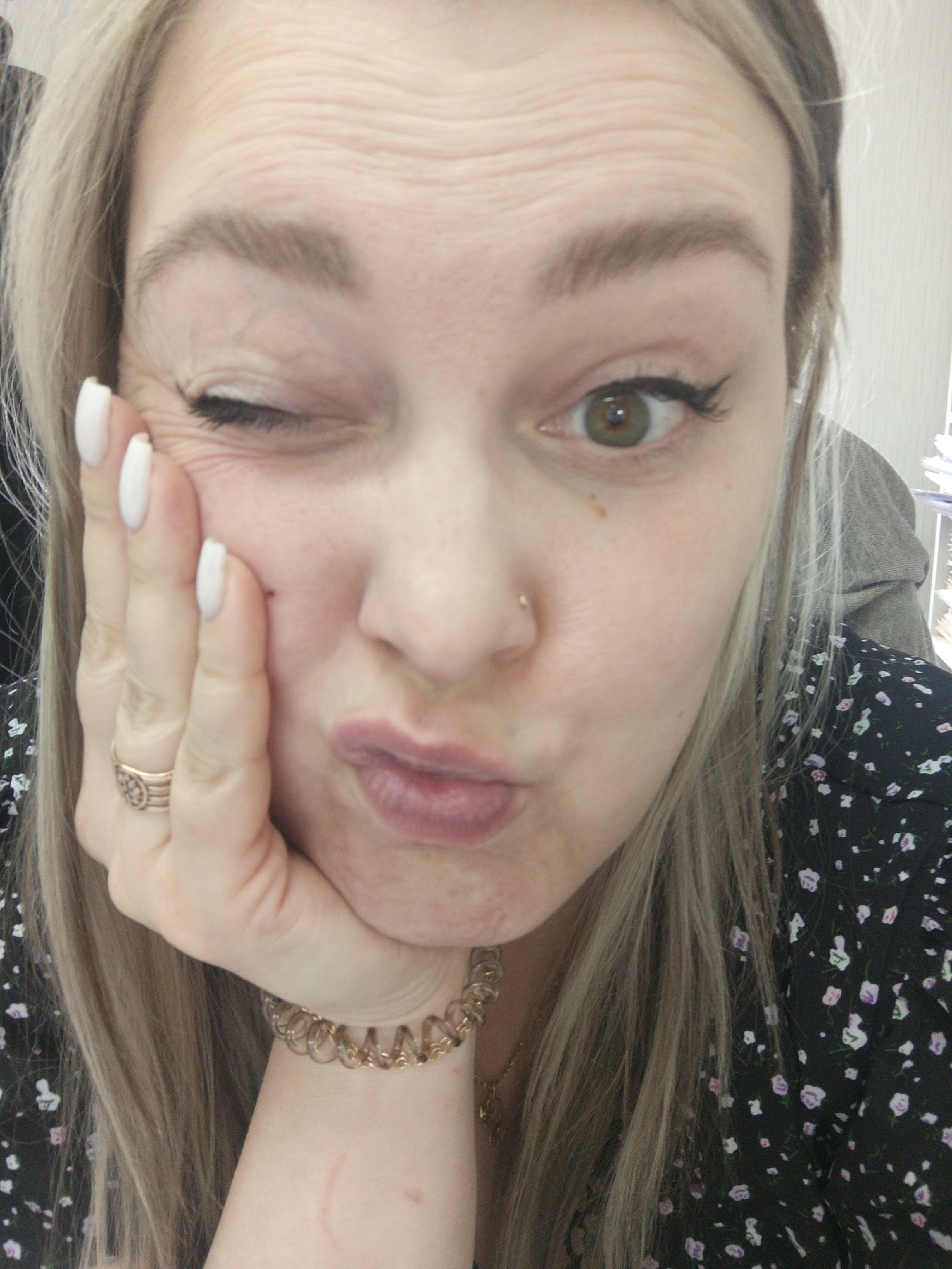 A close-up selfie of a blonde woman with one eye closed in a wink. She has fair skin, light brown eyebrows, and hazel eyes with black eyeliner on the open eye. Her lips are puckered in a slight pout. She has a gold nose ring, a gold ring on her left ring finger, and a gold bracelet on her left wrist. Her left hand is supporting her cheek. She is wearing a dark floral print top. The background is blurry and indistinct. The location, Noviy, Russia, is not discernible from the image.