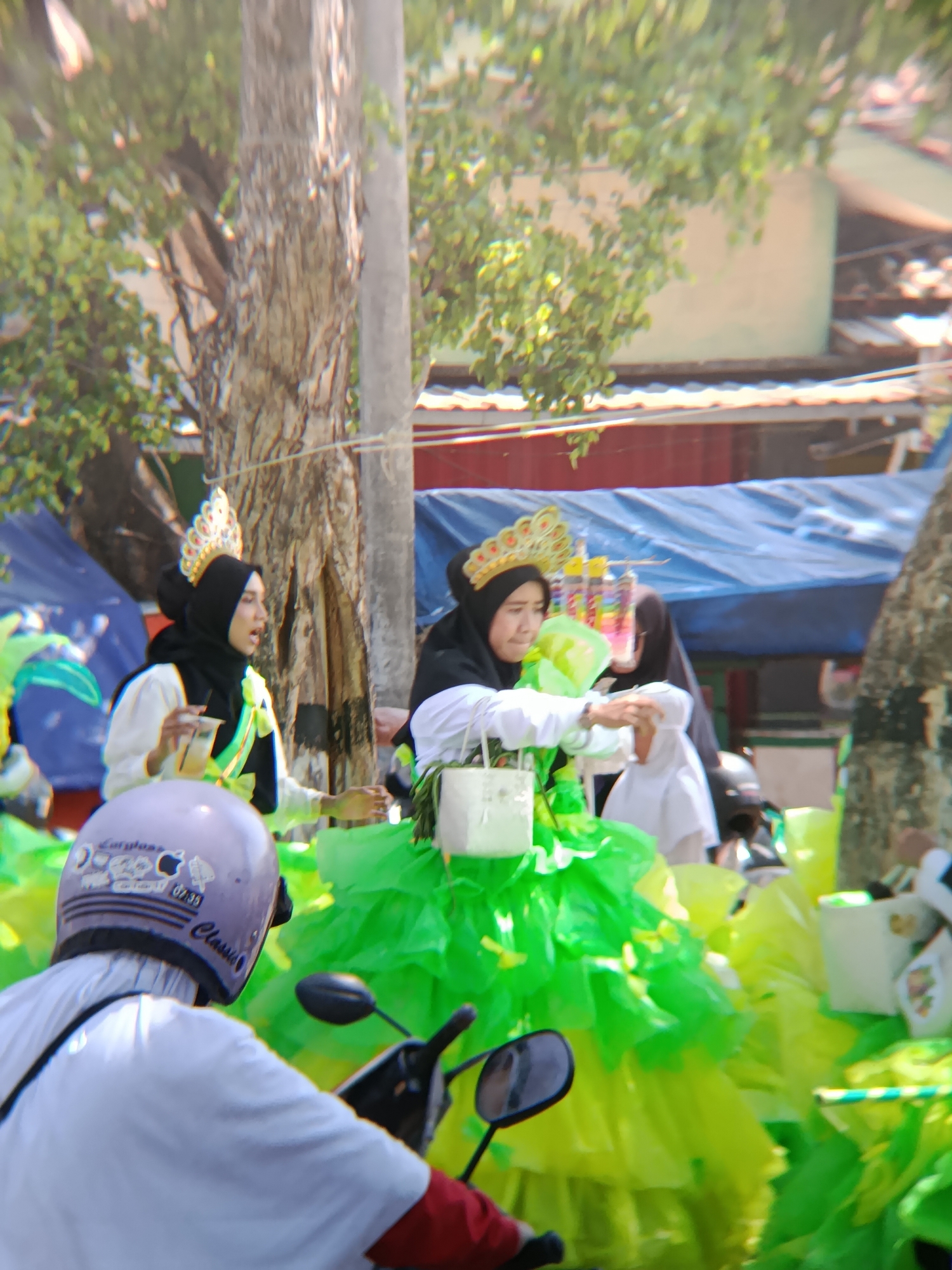 In Desa Kebon Manis, Kecamatan Cilacap Utara, Indonesia, on a sunny day, a parade or festival is underway, indicated by decorated individuals and onlookers. Two women, dressed in traditional Muslim attire with crowns, are prominent. The woman on the left, wearing a black hijab and a white top with a green sash, holds a clear plastic cup and appears to be speaking. She is adorned with a golden crown featuring red and white embellishments. Beside her, another woman, also in a black hijab and white top, is partially visible, holding what appears to be a white purse. Both women are surrounded by vibrant green and yellow fabric, suggesting elaborate costumes or decorations. In the foreground, a person wearing a white shirt and a light purple helmet with stickers, is seated on a motorcycle, looking towards the parade. The background shows tents, trees, and buildings, indicating an outdoor urban or semi-urban setting. The overall atmosphere is festive and celebratory.