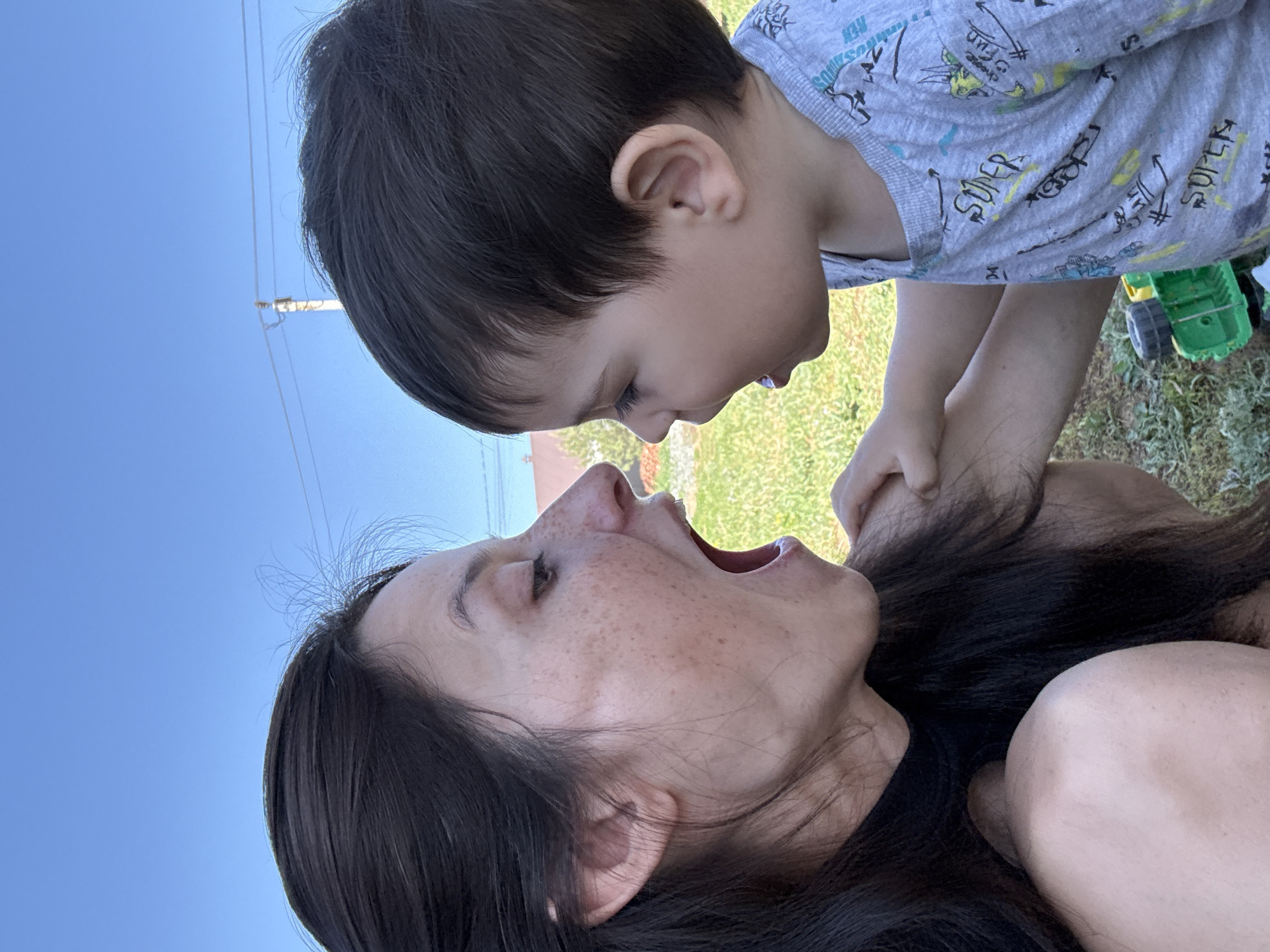 A low-angle, outdoor shot features a woman and a child. The woman lies on her back, with her face tilted up towards the child. Her mouth is open, exposing her teeth and tongue. She has dark hair and visible freckles on her face. The child, above the woman, leans down towards her face. The child has dark hair. They are wearing a grey shirt with various cartoon graphics. A small green toy is visible in the background. The background consists of green grass and a clear blue sky. Power lines are visible. The location is a grassy area in an unknown location.
