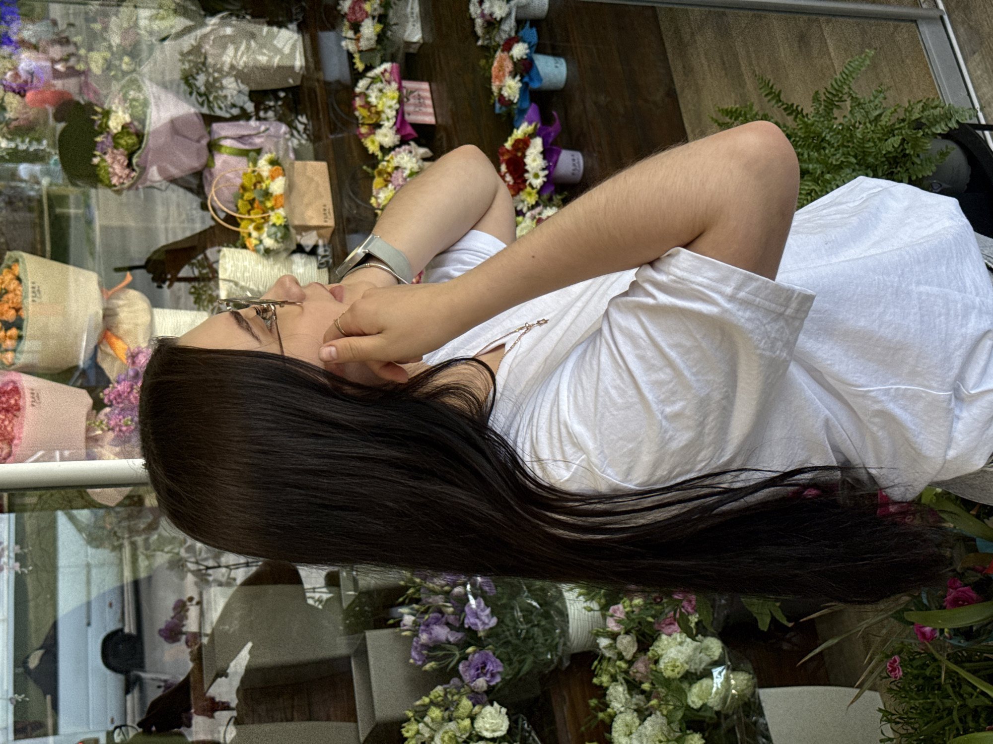 A young woman with long, dark hair and glasses stands in front of a flower shop window. She is wearing a white t-shirt and a silver watch on her left wrist. Her right hand is near her face, touching her ear. The window displays a variety of floral arrangements in different containers. Reflections in the window show a street scene with a white van. The overall environment suggests an outdoor setting in a city.