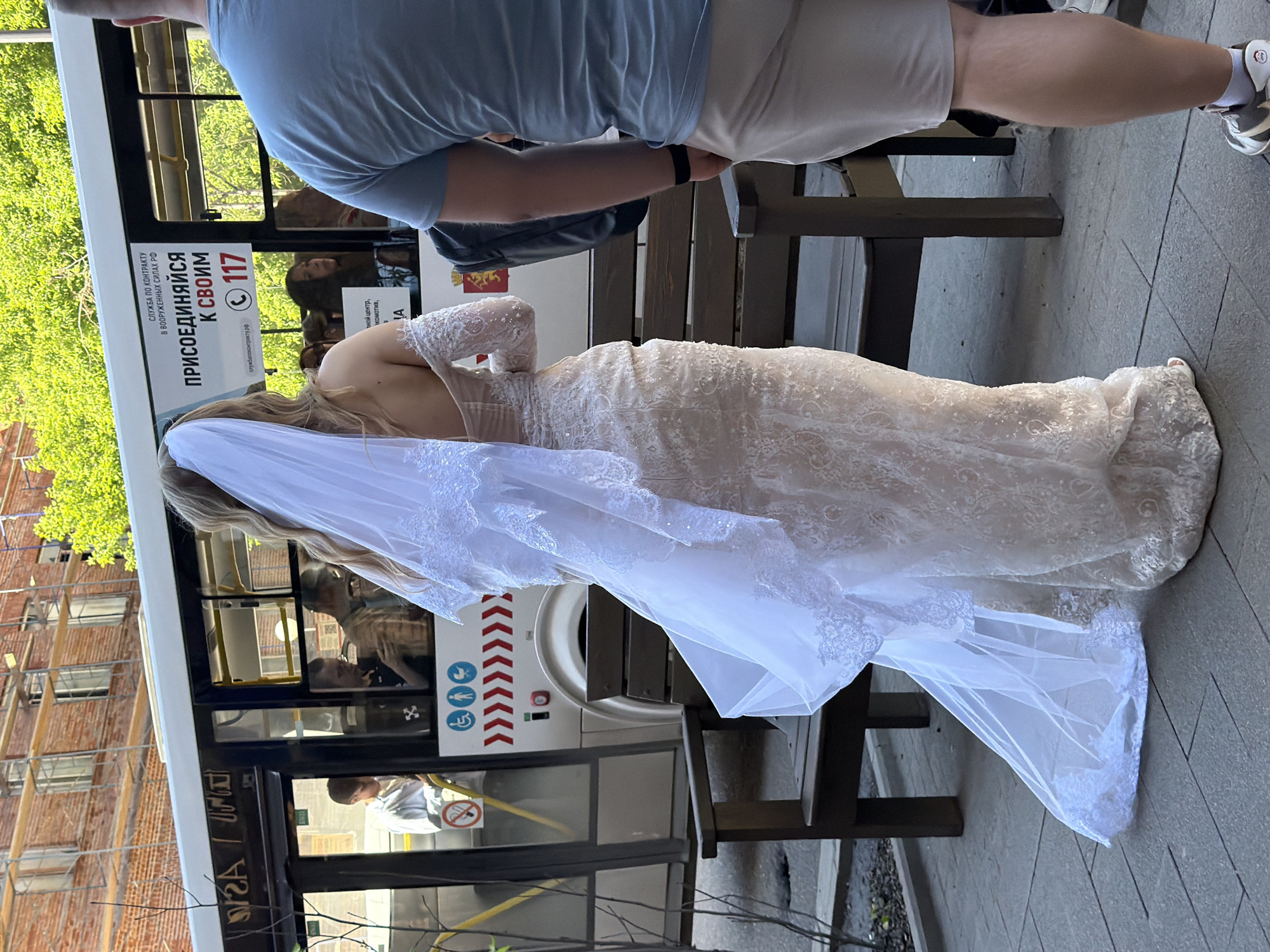 In Krasnoyarsk, Russia, a bride in a white lace wedding dress stands on a sidewalk next to a bus. The bride, with blonde hair and a long veil, is facing away from the camera, her dress adorned with intricate lace. The bus is white with black window frames and displays advertising on its side, including a recruitment message for the armed forces. Inside the bus, several passengers are visible. A wooden bench is positioned on the sidewalk behind the bride. In the background, a brick building is under construction, with scaffolding and exposed framing. The scene is illuminated by daylight, with dappled sunlight suggesting a partly sunny day. A man in a light blue t-shirt and light-colored shorts stands to the right of the bride, also facing away from the camera.