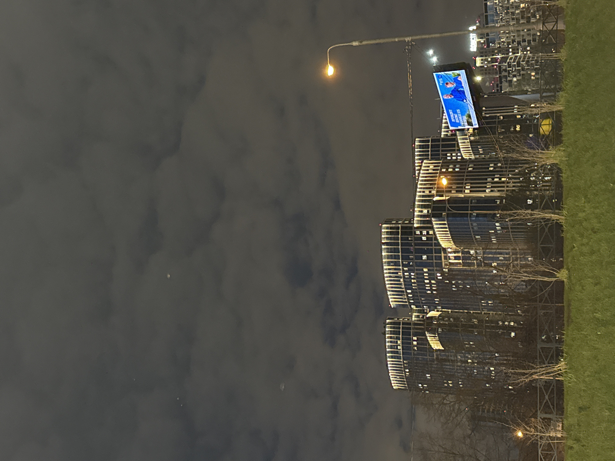 The image, originally rotated 90 degrees counter-clockwise, depicts an urban night scene under a cloudy sky. In its correct orientation, a dark, overcast sky fills the upper left and central areas, with a faint crescent moon discernible amidst the clouds. A street lamp with a warm, orange glow extends into the upper part of the sky.
Below, several modern, multi-story buildings rise, their numerous windows illuminated, suggesting active occupancy. The architectural style appears contemporary, with some facades reflecting ambient light. Text reading "BOHEME CTB" is visible on one building, and "ROST" partially appears on another in the background.
A large, bright digital billboard dominates the mid-right, displaying two men in blue attire, possibly sports figures or public service representatives, against a blue background. Accompanying text appears to be in a non-Latin alphabet, likely Cyrillic. In the far background, behind and above some buildings, a dense area of what appears to be parked cars or carports can be seen.
To the right of the buildings, a strip of vibrant green grass runs along the bottom. Leafless trees or shrubs, indicative of colder seasons, are interspersed between the grass and the building structures. Another street lamp illuminates the lower right foreground. The scene is characterized by artificial lights against the night sky, typical of a city environment in Khimki, Russia.