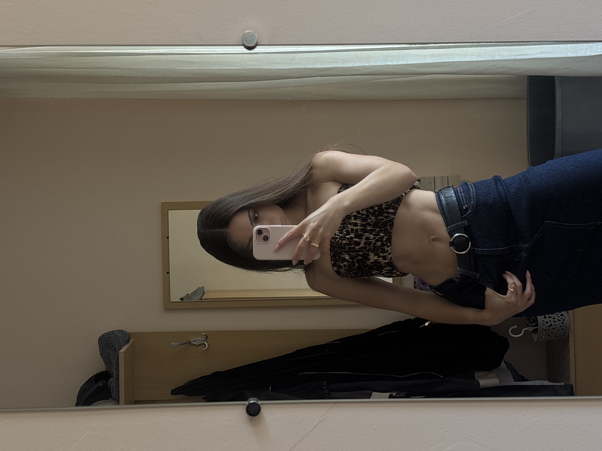 A young woman is taking a mirror selfie in what appears to be a bedroom or dressing area, possibly in Varna, Bulgaria. She is wearing a leopard print bandeau top and dark wash denim jeans. Her left hand is holding a pink smartphone, and her right hand is casually placed in her jeans pocket. She is wearing rings on both hands. The background includes a wardrobe with hanging clothes, a shelf with some items, and a large mirror that reflects her and the surrounding space. A sheer curtain is visible on the right side of the frame.