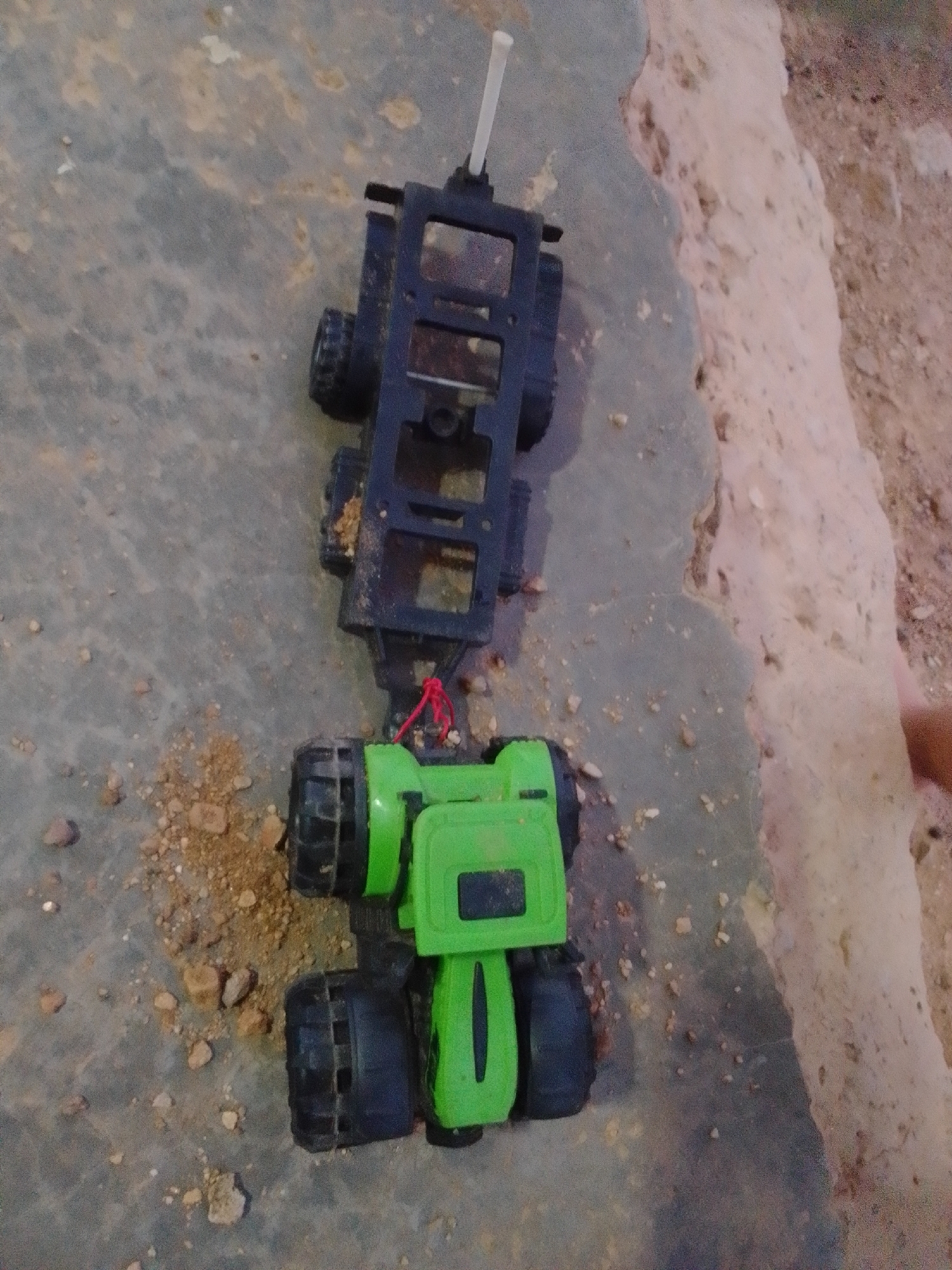 The image shows a toy truck on a dirt and concrete surface. The toy truck is black and green and is connected by a red string. The background is a rough, textured surface that appears to be a mix of dirt, rocks, and possibly concrete. There are scattered pebbles and dirt on the ground around the toy truck. The image is taken from a top-down perspective. There are no people visible in the image. The location of La Tagua, Venezuela is not discernible from the image itself.