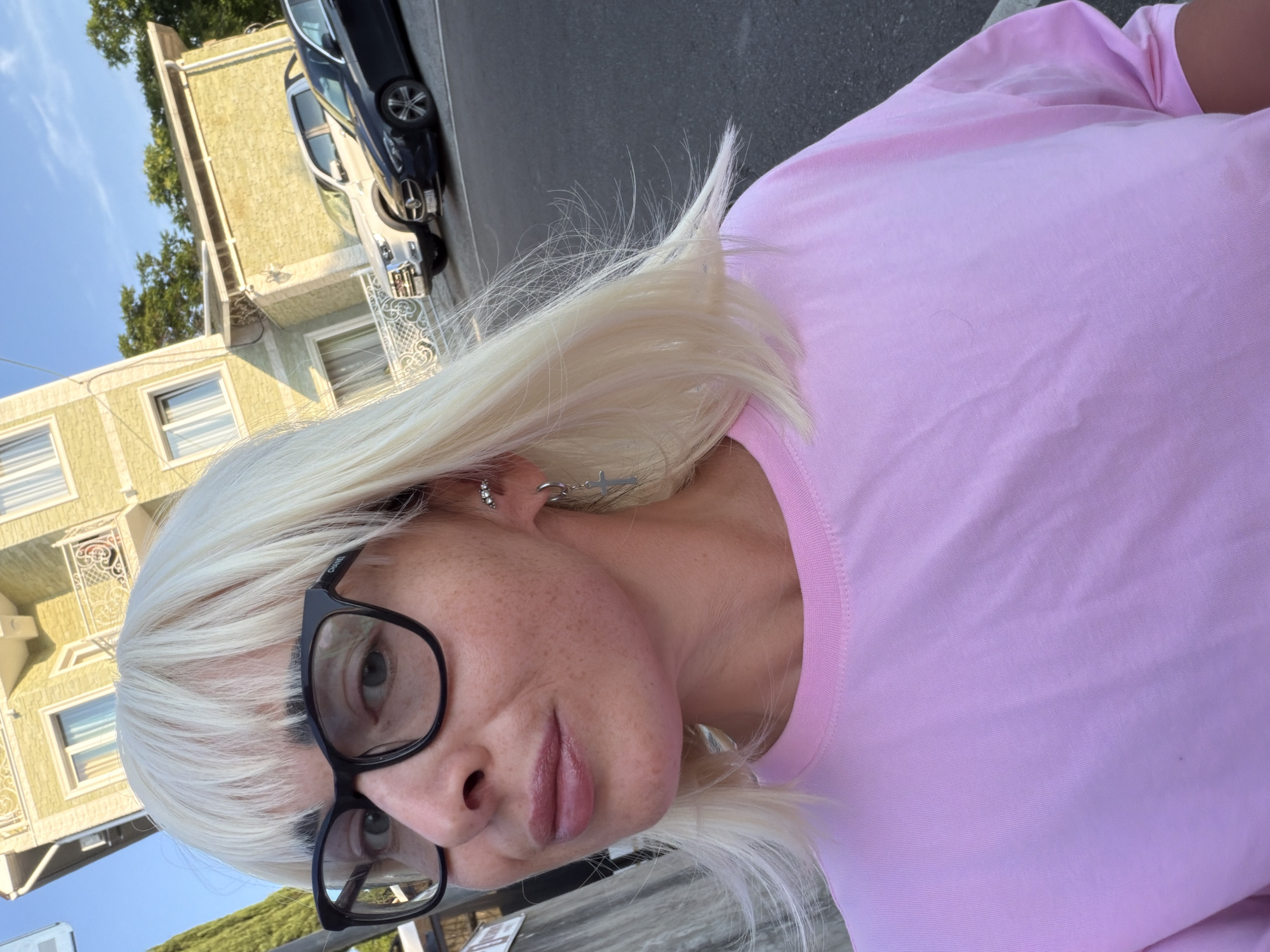 A selfie taken outdoors in Sochi, Russia, features a woman with platinum blonde hair and bangs. She is wearing black-rimmed glasses and a light pink t-shirt. A silver earring with a cross charm is visible on her left ear. The background includes a building with a yellow facade and balconies, trees, and parked cars, one of which appears to be a dark-colored sedan and another a white pickup truck. The scene suggests a sunny day with clear skies.