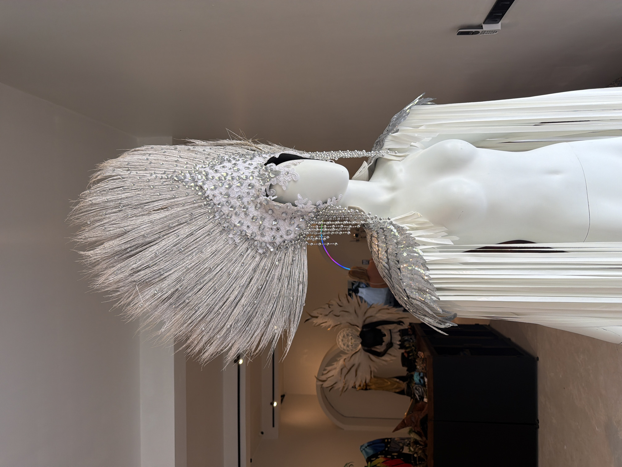 In Ko Samui District, Thailand, a white mannequin is displayed wearing an elaborate silver headdress adorned with feathers and intricate beadwork. The headdress fans outwards dramatically and is embellished with cascading silver fringe. The mannequin also wears a cape or shoulder piece with similar silver detailing and long white fringe. The background suggests an indoor retail space, possibly a costume shop, with shelves and other decorative items visible. Another mannequin with large white wings is seen in the background, along with a glimpse of a person. The overall impression is one of a festive or performance-oriented costume on display.