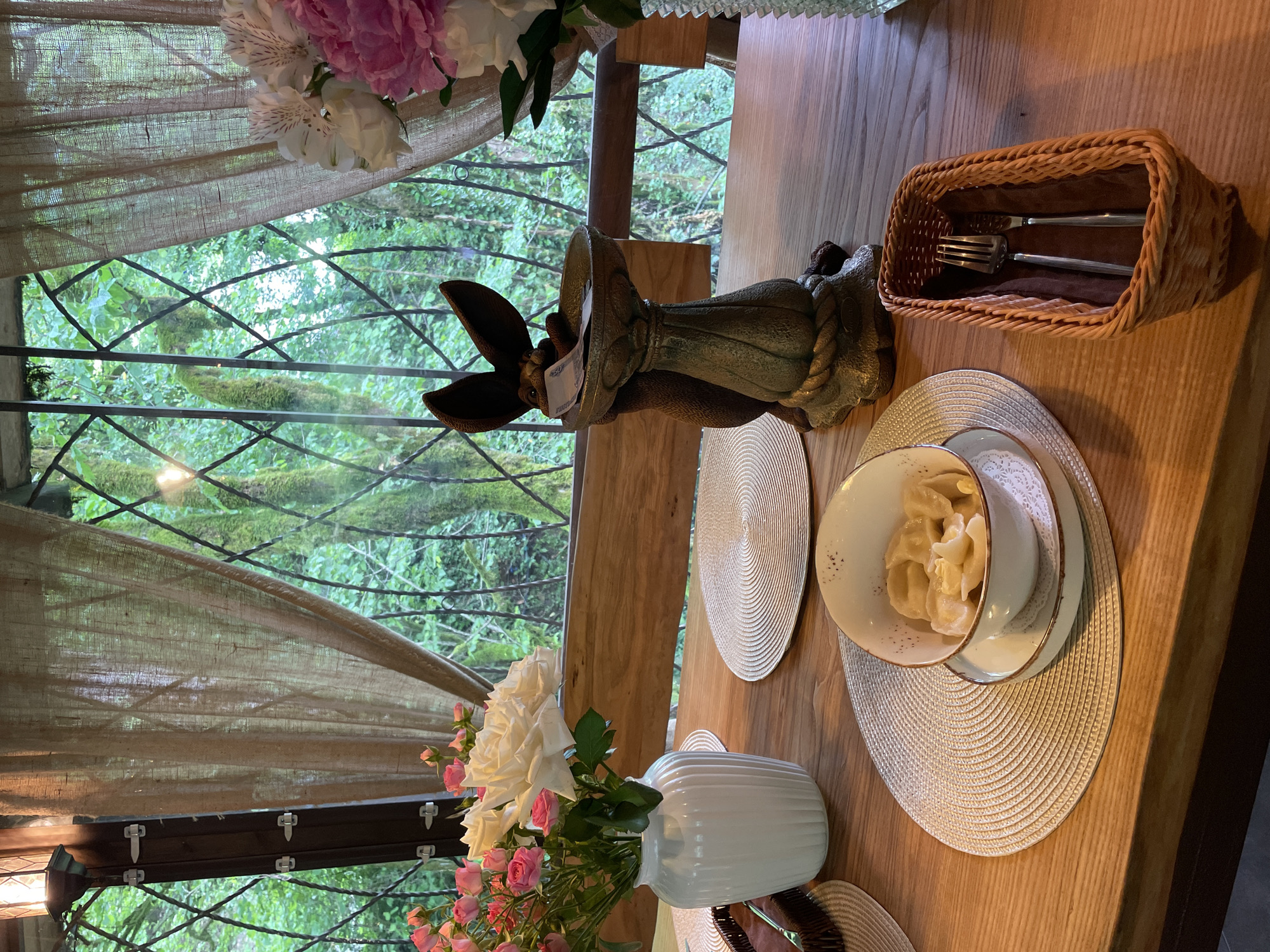 The image shows a dining table set for a meal. A decorative bunny sculpture stands near a window overlooking a lush green forest. A wicker basket with silverware sits on the table, along with a plate setting consisting of a bowl, plate, and woven placemat. A vase filled with white and pink roses is also on the table. The window is covered with a sheer curtain. This scene is likely located in Solokh-Aul, Russia, suggested by the presence of the forest setting and the dining setup.