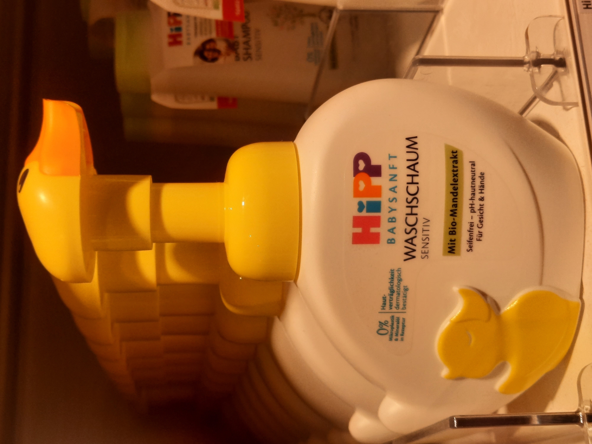 The image shows a row of white plastic bottles with yellow duck-shaped dispensers, likely baby wash products, displayed on a shelf in a retail environment. The bottles have branding that includes the "HiPP" logo and text in German, such as "BABYSANFT WASCHSCHAUM SENSITIV". One bottle in the foreground is in focus, showing the label clearly. The background is blurred, with other products and shelves visible. The lighting suggests an indoor setting, possibly a supermarket or drugstore. The image does not provide specific clues about its exact location within Rosenheim, Germany.