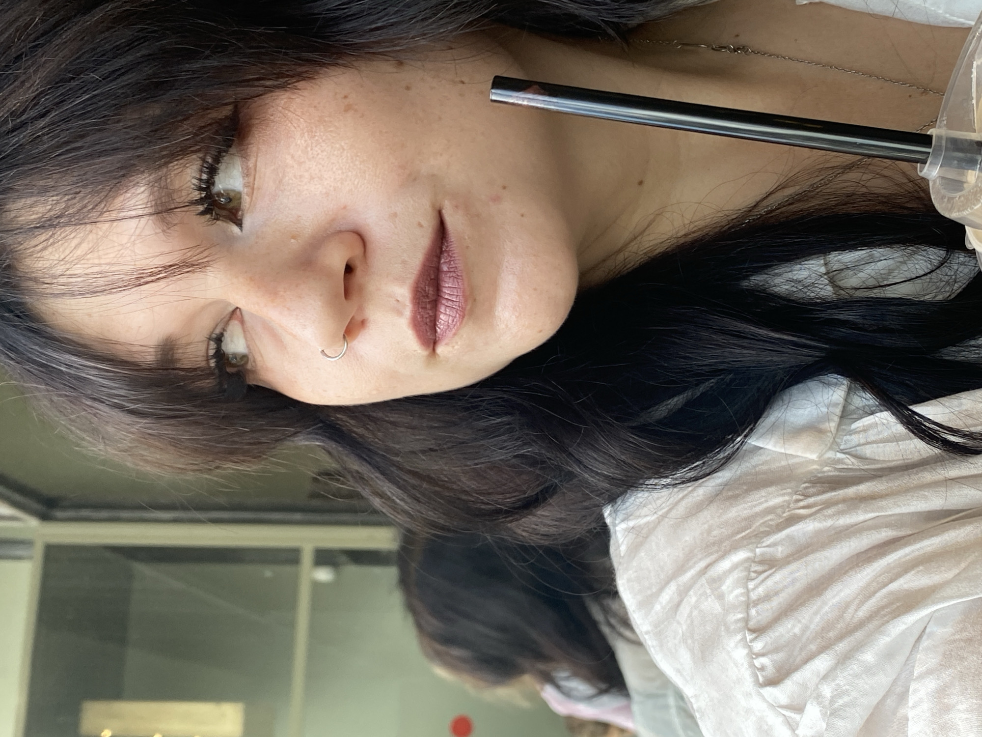 A medium close-up shot shows a young woman with long, dark, wavy hair and light brown eyes, looking to her left. She has a silver hoop nose ring and wears a light-colored, possibly white or cream, collared shirt. Her makeup includes dark eyeliner and a muted berry-colored lipstick. A dark, slender straw protrudes from a clear plastic cup held by the woman. The background is out of focus, showing a reflection of what appears to be another person and a window with a greenish tint. The lighting suggests an indoor setting, possibly a cafe or restaurant.