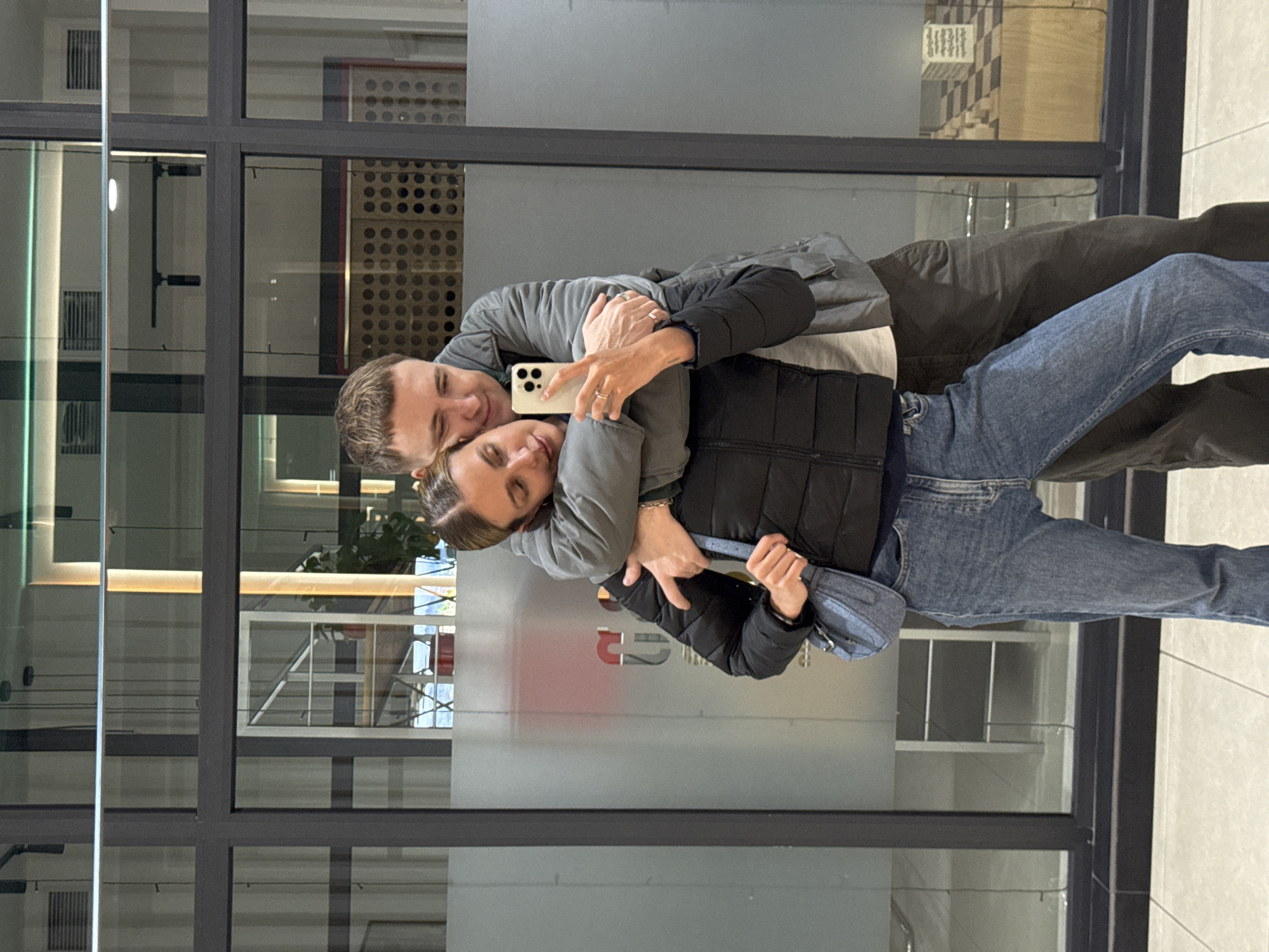 A man and woman stand close together, posing for a selfie with a smartphone. They are dressed in casual clothing, the man in a gray jacket and brown pants, and the woman in a black vest, denim jeans, and a blue fanny pack. They are positioned in front of a glass window that reflects interior architectural elements and daylight. The location is in Tbilisi, Georgia.
