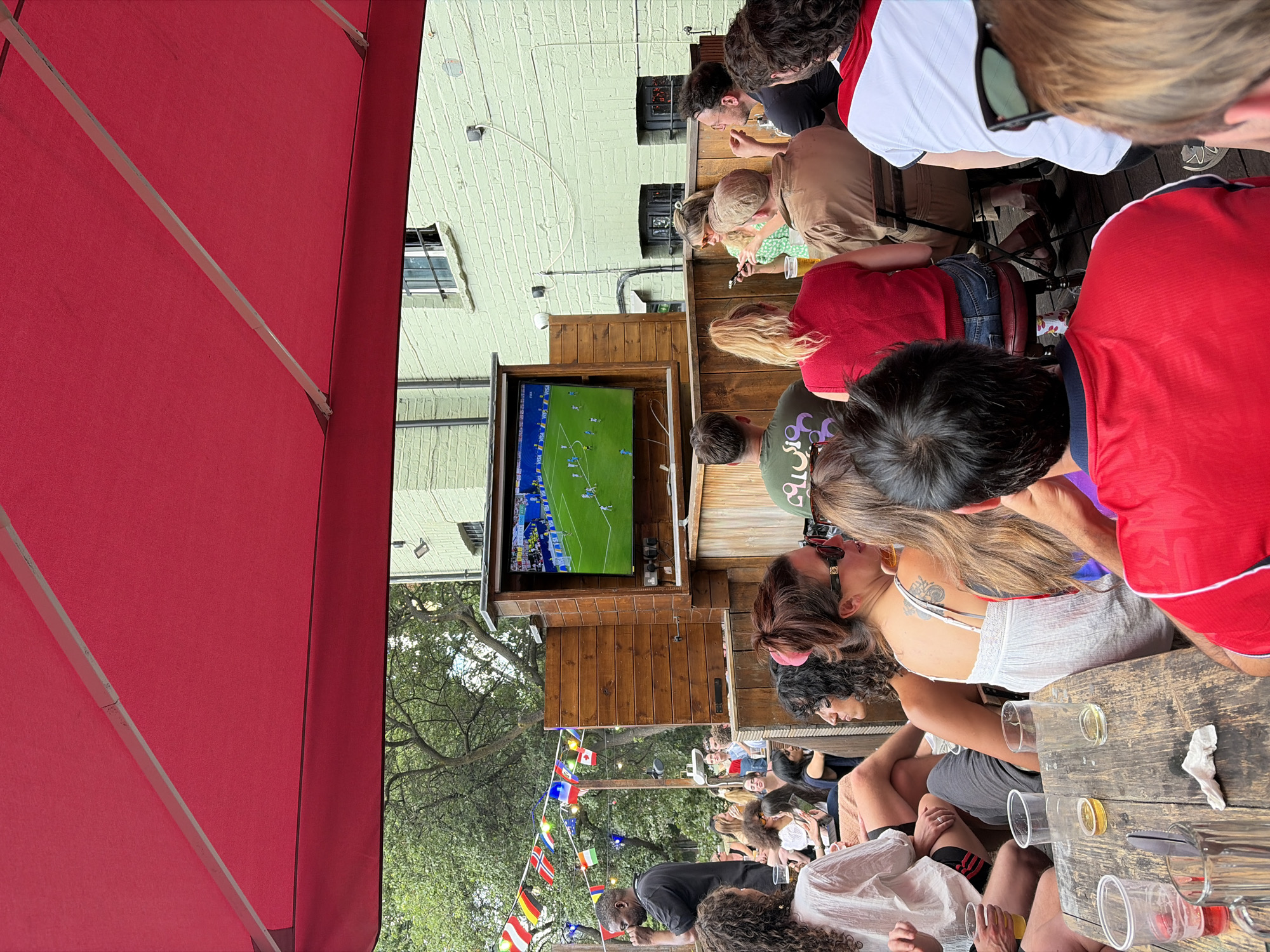 The image captures a lively outdoor gathering at what appears to be a bar or pub, with a group of people watching a soccer match on a television screen. The scene is set on a wooden deck under a red awning. The people, dressed casually, are seated and standing around, some facing the screen, others interacting amongst themselves. A woman in a white top is kissing a man, indicating a social and relaxed atmosphere.
The television displays a soccer game, suggesting the event is a live broadcast of a match. Numerous small flags, including those of Germany, Ireland, and Belgium, are strung across the area, hinting at an international sporting event or a venue celebrating diverse nationalities. The presence of beer glasses on wooden tables, along with the casual attire of the patrons, points towards a leisurely afternoon or early evening. The lighting suggests it is daytime, possibly late afternoon or early evening, with some shadows cast by the trees visible in the background. The building behind the gathering has a light-colored brick facade. The overall mood appears to be jovial and engaged, with people enjoying themselves amidst the game and each other's company. There is no visible text in the image apart from what is on the soccer game screen.