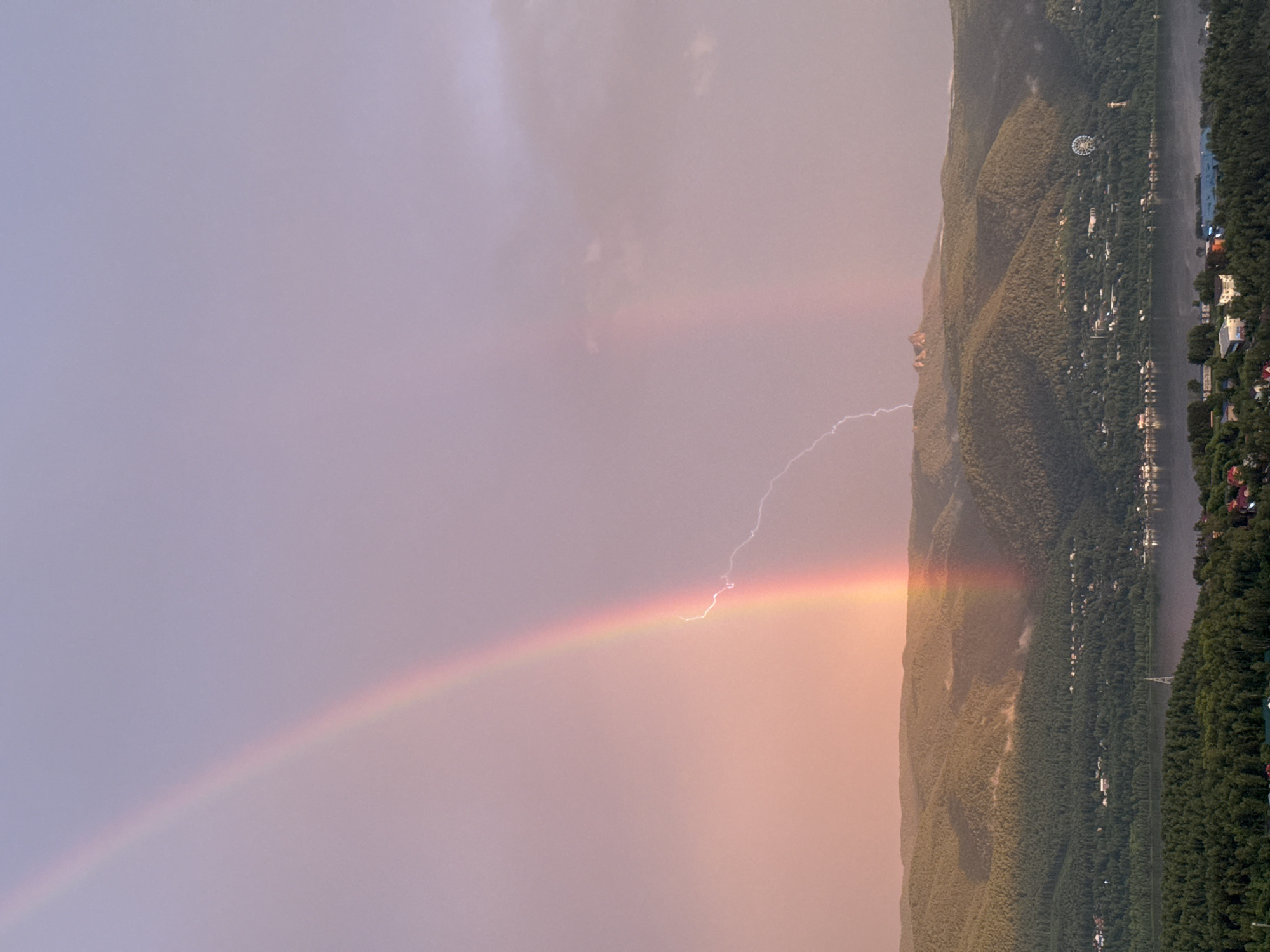 The image captures a dramatic natural phenomenon over a scenic landscape that appears to be the city of Krasnoyarsk, Russia. A vibrant rainbow arcs across a soft, pastel sky, illuminated by the warm hues of what seems to be either sunrise or sunset. Intersecting the rainbow, a bright bolt of lightning streaks downwards, creating a striking contrast between the serene colors of the rainbow and the raw power of the storm.
The foreground is dominated by rolling, tree-covered hills that descend towards a wide body of water, likely the Yenisei River. Nestled within the foothills and along the riverbanks are scattered buildings, indicating a populated area within this natural setting. A Ferris wheel is visible on one of the hills, a common landmark in urban recreational areas. In the distance, the jagged peaks of mountains can be seen, adding to the majestic backdrop. The overall impression is a fleeting moment of intense weather activity occurring over a beautiful and inhabited natural environment.