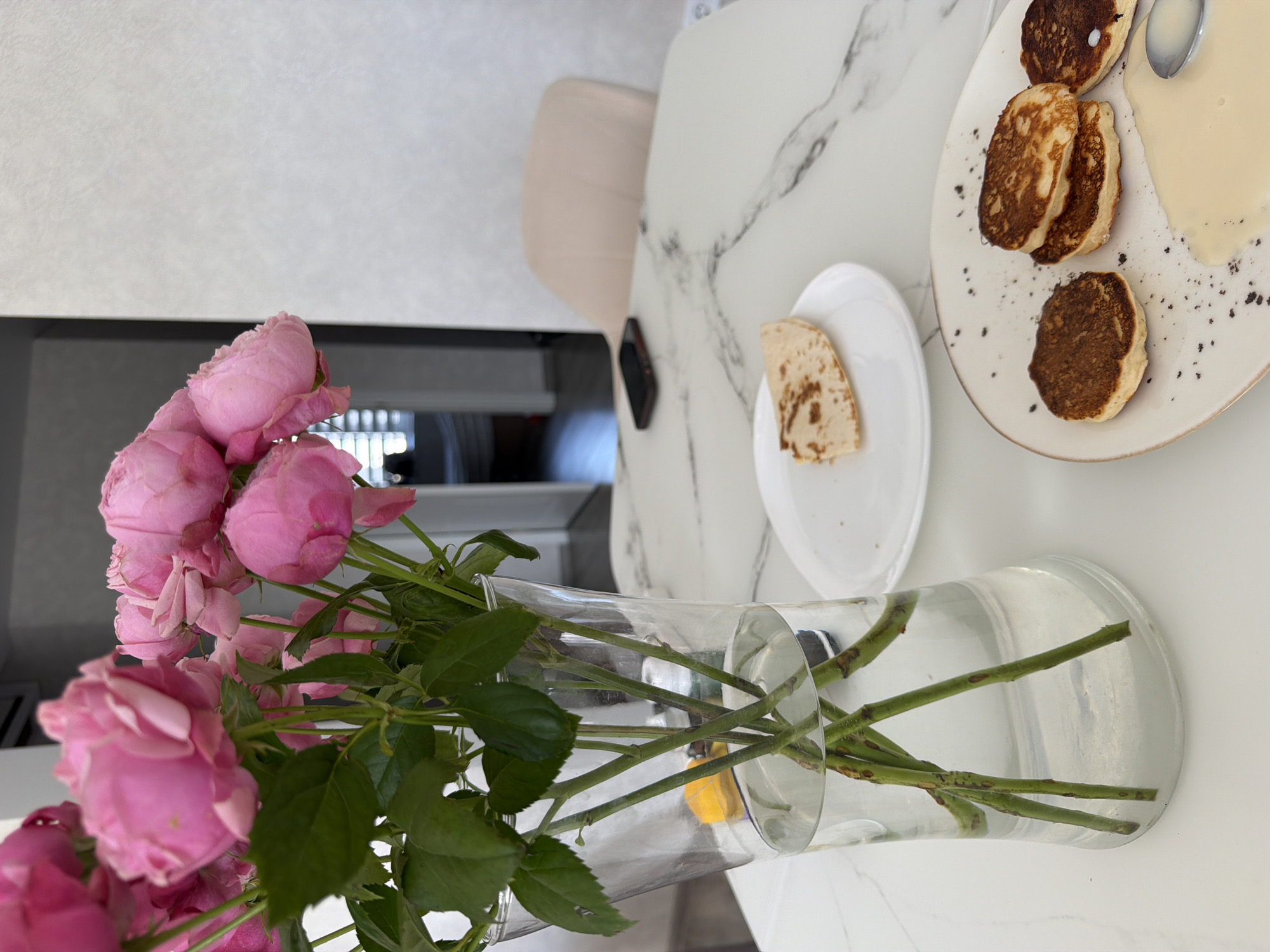 A floral arrangement of pink roses in a clear glass vase is positioned on a white marble-patterned table. To the right of the vase, a white plate holds a small, folded piece of flatbread. Further to the right, another plate contains several small, golden-brown pancakes, some stacked, with a pool of light-colored sauce and a spoon resting in it. In the background, a light-colored chair is visible, and a smartphone lies on the table near the chair. The scene appears to be indoors, possibly a dining area in Malinovka, Russia.