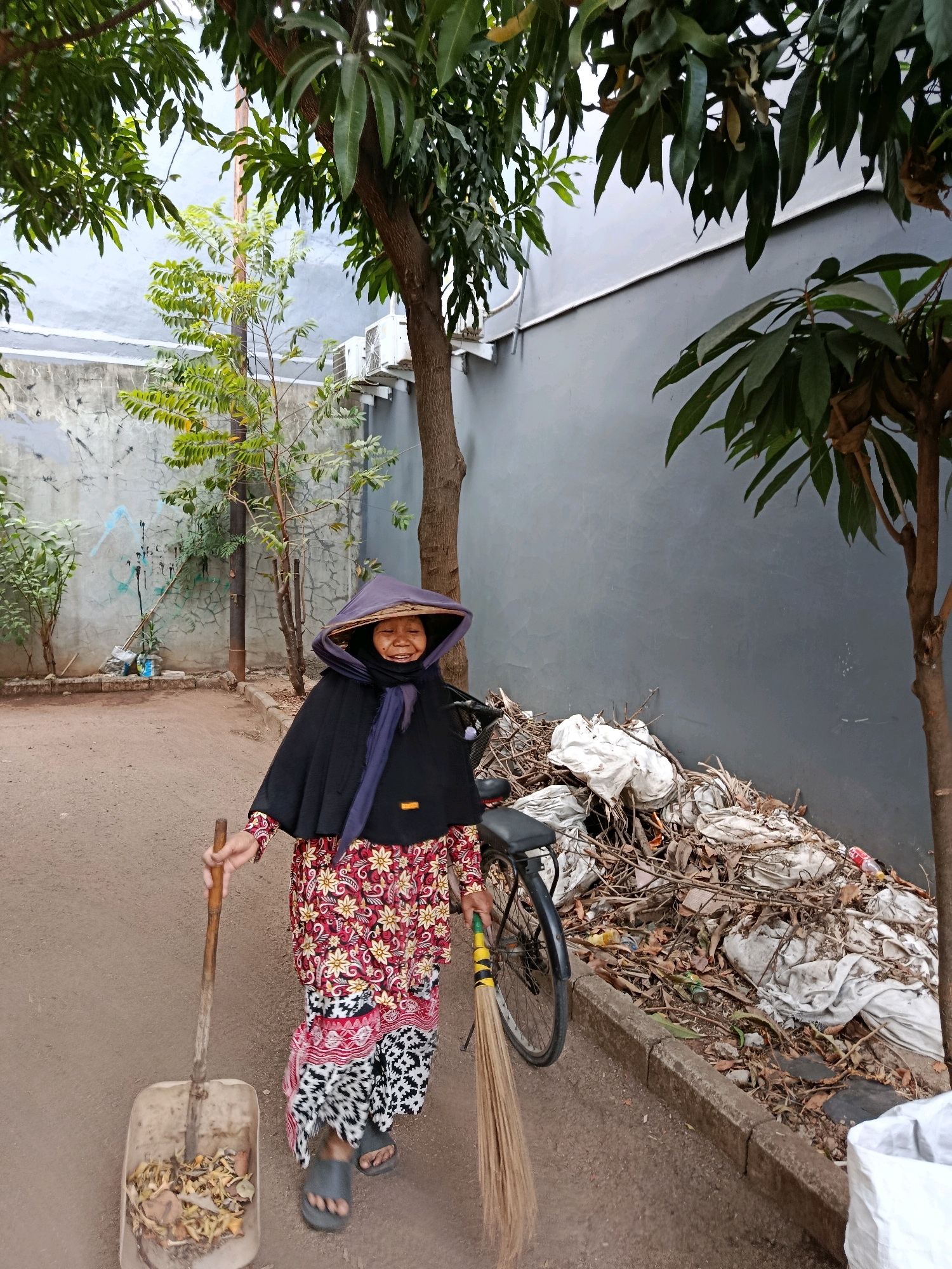 An elderly woman wearing a traditional conical hat and a black cape over a patterned red and white dress stands in an outdoor area. She is holding a shovel in her left hand and a broom in her right hand. The shovel is positioned over a dustpan filled with leaves and debris. To her right, a pile of trash bags and branches is visible next to a bicycle. The background consists of a grey wall, a concrete wall with graffiti, and trees with green leaves. The ground is dirt and asphalt. The scene appears to be in a residential or industrial area. This image was taken in the city of Bekasi, Indonesia.