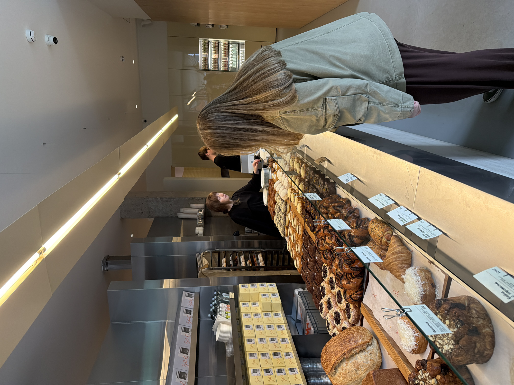 The image shows the interior of a bakery, possibly in Moscow, Russia based on the Cyrillic script on the price tags. A woman with long, light brown hair is standing at the pastry display case, which is filled with a variety of breads, pastries, and cakes. Behind the counter, a person in a black shirt appears to be working. Boxes and supplies are stored behind the counter. The overall lighting is bright and the setting appears modern and clean.