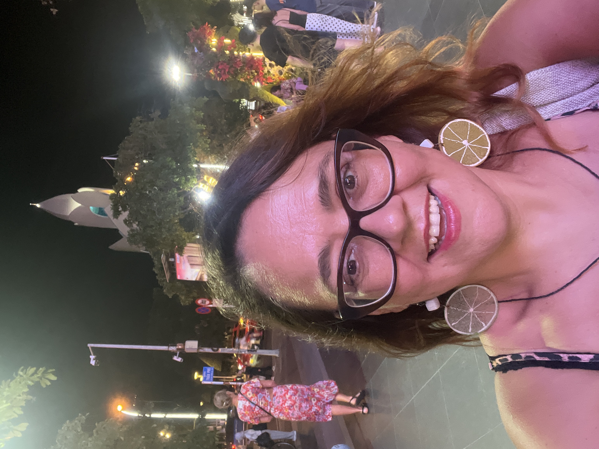 This is a selfie taken at night on a street in Nha Trang Ward, Vietnam. The foreground features a woman with brown hair, wearing glasses, and large circular earrings that resemble sliced citrus fruit. She is smiling and looking at the camera. She is wearing a patterned top. In the background, a woman in a floral dress walks away from the camera. Other people and traffic are visible further down the street, illuminated by streetlights and festive decorations. A distinctive white building with a pointed, multi-layered roof is visible in the distance, lit up at night. The scene appears to be a lively urban environment.