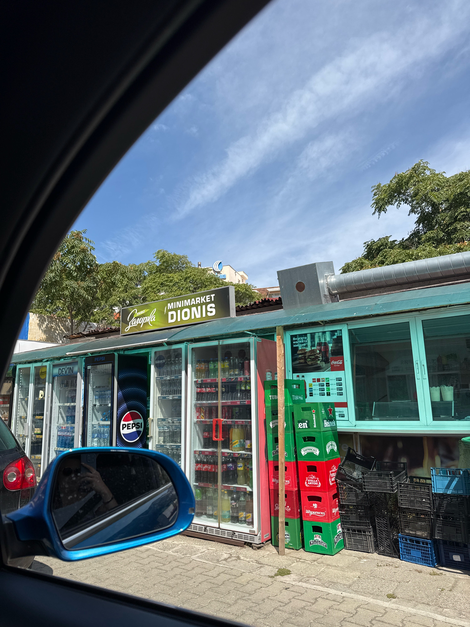This image depicts a minimarket named "Dionis" in Sozopol, Bulgaria. The storefront features large glass refrigerators stocked with beverages, including Pepsi and Coca-Cola. Stacks of red and green crates, some labeled with beer brands like Stella Artois and Heineken, are piled outside the establishment. A blue car mirror is visible in the foreground, reflecting a person wearing sunglasses. The minimarket appears to be situated along a street with a paved sidewalk. The sky above is blue with scattered white clouds, and trees are visible in the background.