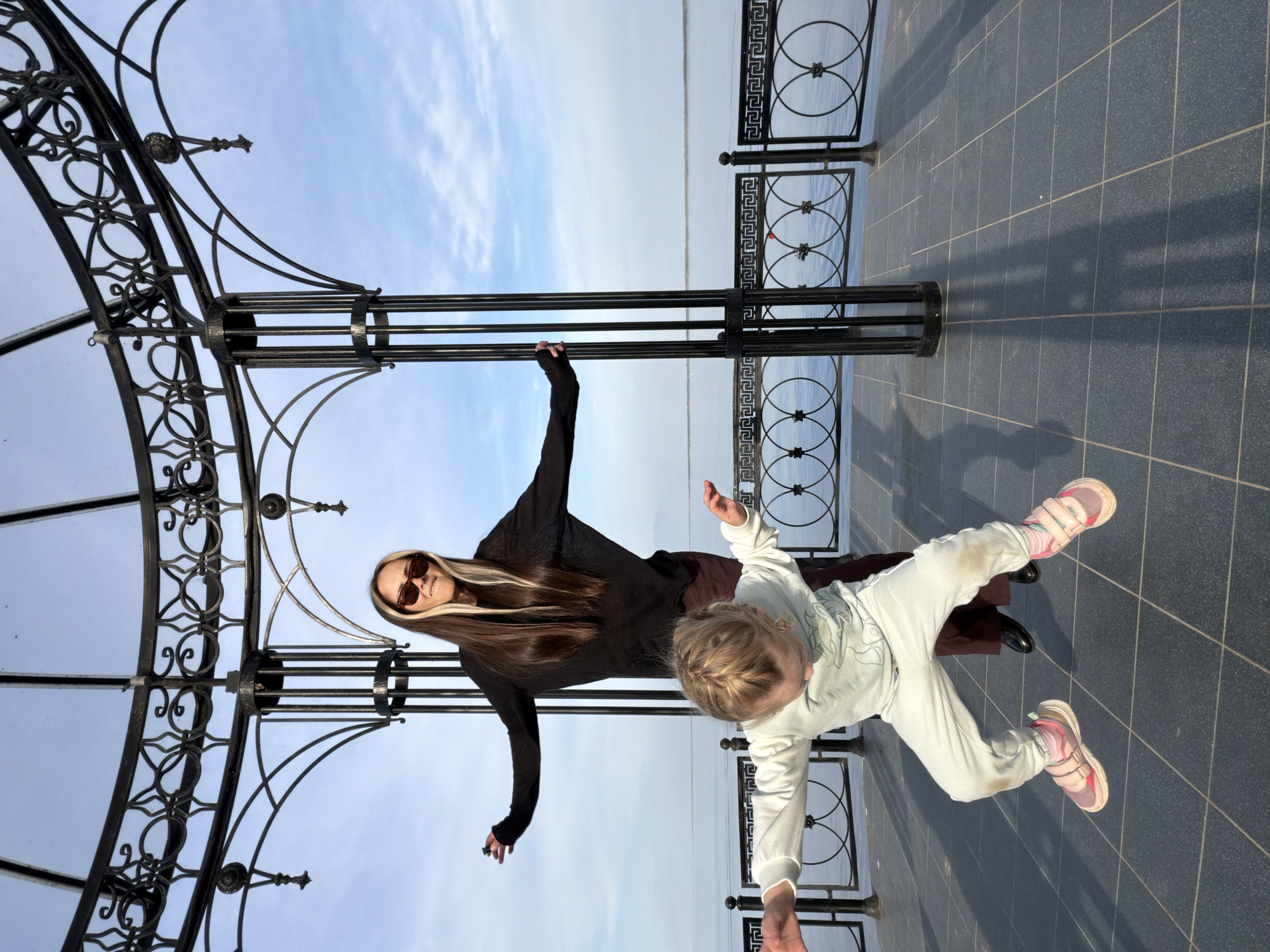 The image shows a woman and a young girl posing near a metal gazebo and railing in Kamyshin, Russia. The woman is wearing a dark long-sleeved shirt, long dark skirt, and sunglasses. Her hair is dark with blonde streaks. The young girl is wearing a white sweatsuit and pink sneakers. They are both holding their arms out to the sides. The gazebo and railing are black metal. The ground is tiled with dark gray square tiles. In the background is a body of water, with a pale blue sky above.