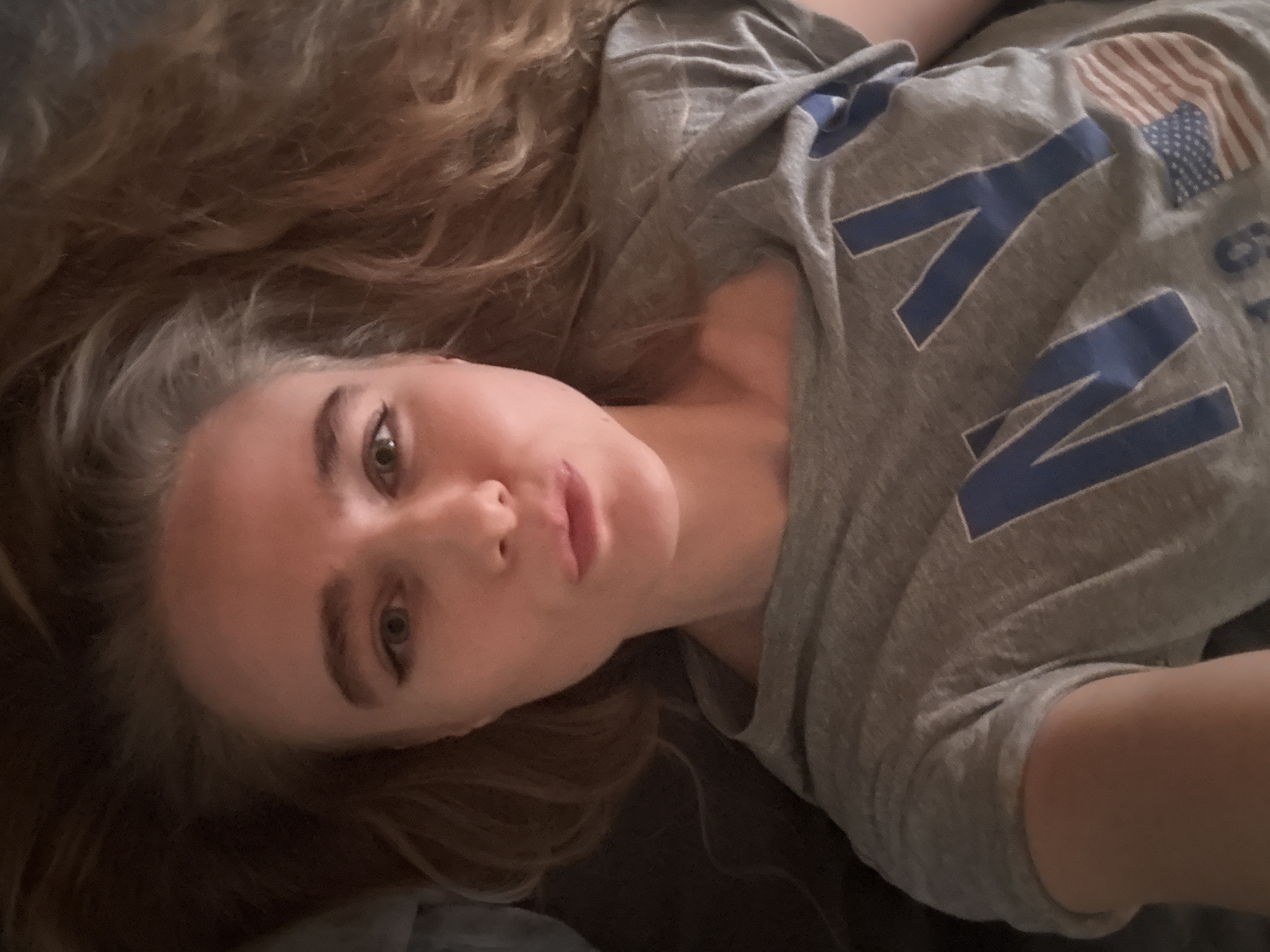 A young woman with long, light brown hair is lying on her back, looking at the camera. She has fair skin, light eyes, and her hair is spread around her head. She is wearing a grey t-shirt with "NY" printed in blue on it, along with a faded American flag design. The background is dark and indistinct. The location is unknown.