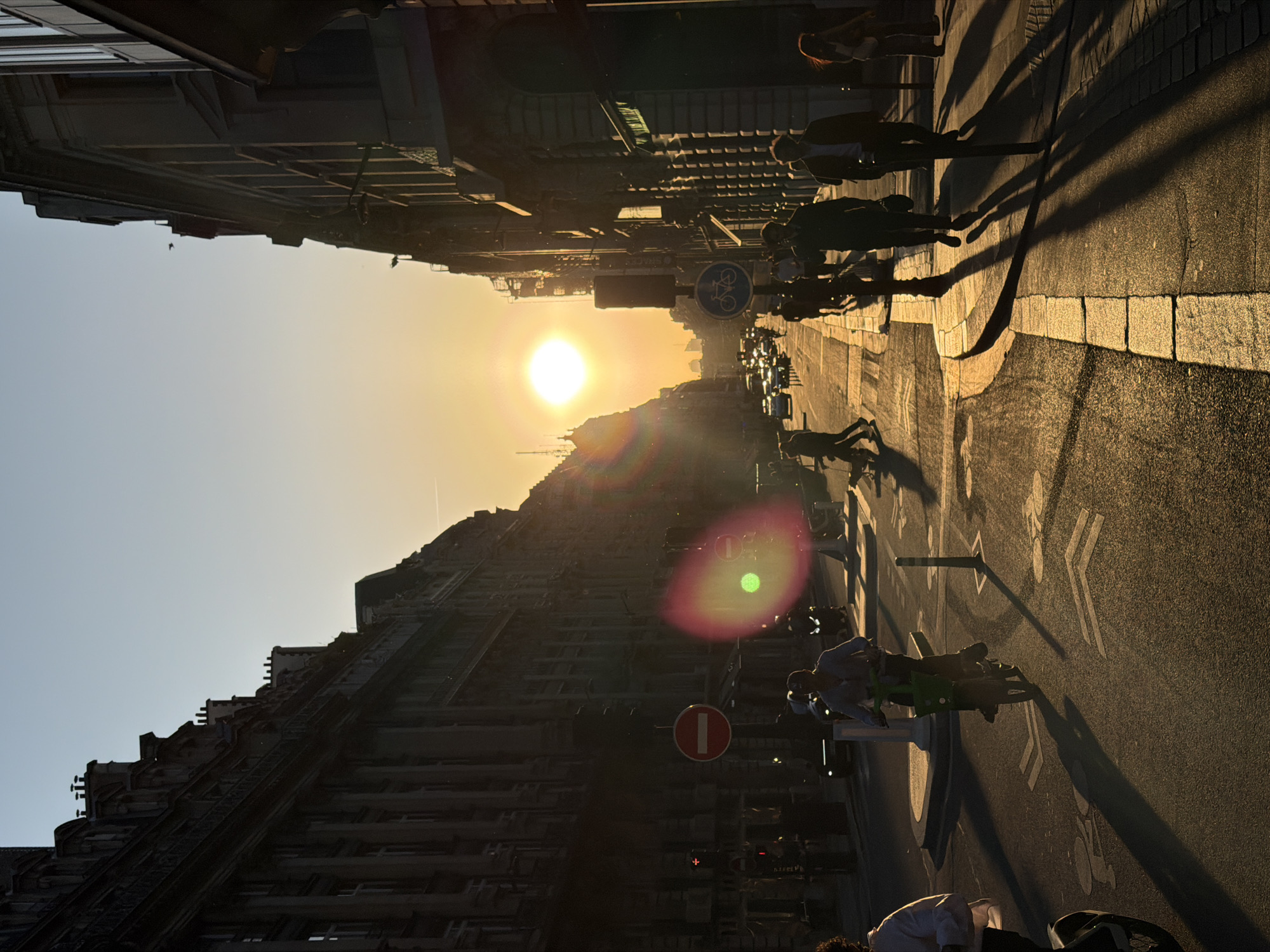 This image captures a street scene in Paris, France, at what appears to be sunrise or sunset. The sun is positioned high in the sky, casting long shadows of pedestrians walking on the right side of the street. The architecture is distinctly Parisian, with tall buildings lining both sides of the road. There are street signs visible, including a bicycle lane sign and a "do not enter" sign. Cars are parked along the street, and a person is riding a green scooter or bike on the bike lane. The road markings indicate the presence of a dedicated bike lane. The overall atmosphere is one of urban activity during a time of day when sunlight is prominent.