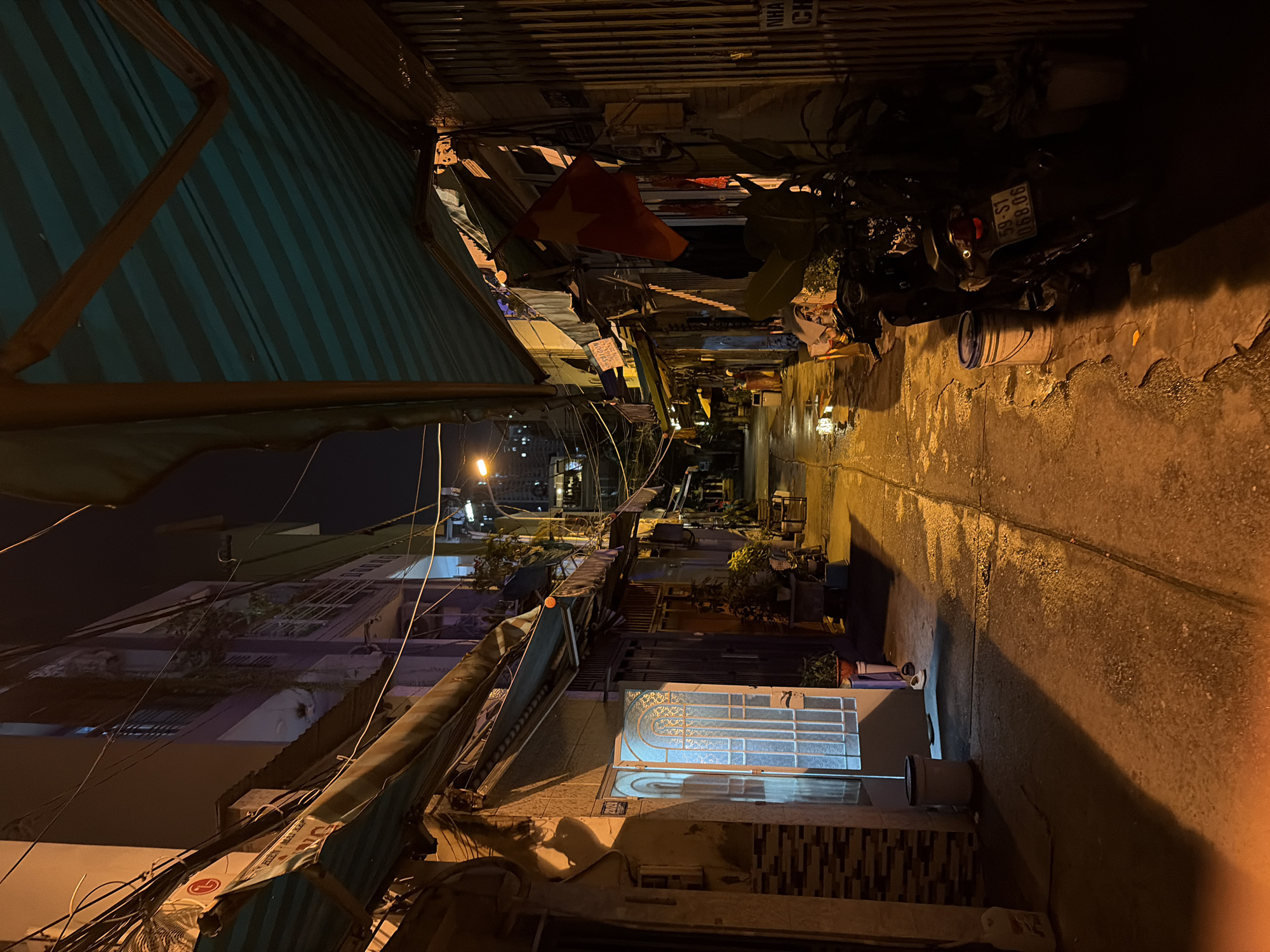 The image captures a narrow urban alleyway at night, illuminated by artificial light sources that cast a warm, yellowish glow on the wet pavement. The scene is viewed from a slightly elevated vantage point, looking down the length of the alley.
On the left side, a large awning with green and blue stripes extends over the space, supported by wooden or metal beams. Beyond it, multi-story buildings line the street, characterized by windows, balconies, and a dense network of overhead electrical wires. A distant streetlight or building light is visible further down the alley.
The right side of the alley is also flanked by buildings, with varied architectural details including tiled sections and plain walls. A prominent red Vietnamese flag with a yellow star hangs from one of these structures. Several motorbikes are parked along the curb, and one clearly displays a Vietnamese license plate: "59-S1 068.06." A white bucket is situated near the parked vehicles. The ground on this side is visibly wet, reflecting the overhead lights and indicating recent rain or puddles. A building features a distinctive door or window with an intricate, glowing white pattern.
Visible text includes the motorbike license plate "59-S1 068.06" (confirming a Vietnamese location, specifically Ho Chi Minh City). Other partial text appears on signs and awnings; a fragment resembling the "LG" logo is visible on a torn awning, and "NHA CH..." is partially seen on a sign near the Vietnamese flag, possibly indicating a type of establishment. No people or overt activities are visible, lending a quiet, atmospheric feel to this common urban scene in Thủ Đức, Vietnam.