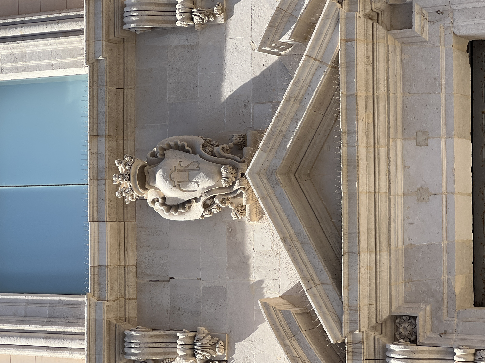 This is a close-up, eye-level shot of a stone architectural detail above an entrance in Syracuse, Italy. The detail features a shield-like crest with the letters "IHS" inscribed within, topped by a crown. The crest is flanked by ornate carvings and supported by decorative brackets. The stonework appears aged and weathered. Above this, a large blue window is visible, and along the ledge of the window and another horizontal architectural element, there are rows of metal bird spikes. The scene is illuminated by natural daylight, casting shadows on the stone surfaces. The overall impression is one of historical grandeur and meticulous craftsmanship.