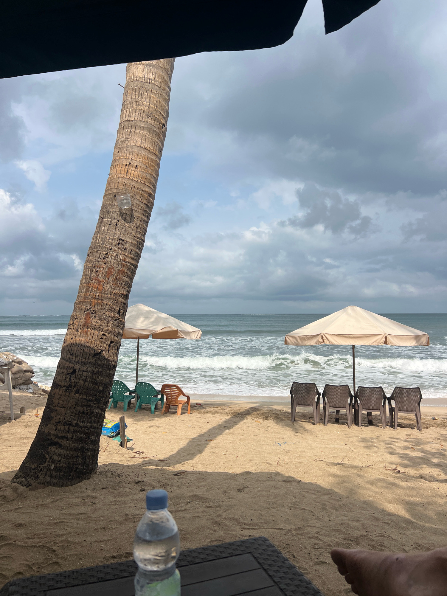 This image depicts a beach scene in Kuta, Indonesia, under a cloudy sky. A large palm tree trunk occupies the left foreground, casting a shadow on the sandy ground. In the middle ground, two beach umbrellas are set up, one with a cream-colored canopy supported by a dark pole, and another, partially visible, to its left. Beneath the left umbrella, three green plastic chairs and one orange plastic chair are arranged. Beneath the right umbrella, four dark-colored plastic chairs are lined up facing the ocean. The ocean waves are visible in the background, crashing onto the shore. In the immediate foreground, a dark table with a woven texture is visible, with a bottle of water placed on it. A person's foot is also visible on the right side of the frame, resting on the table. The overall atmosphere is overcast, suggesting a potentially rainy or storm-prone day.