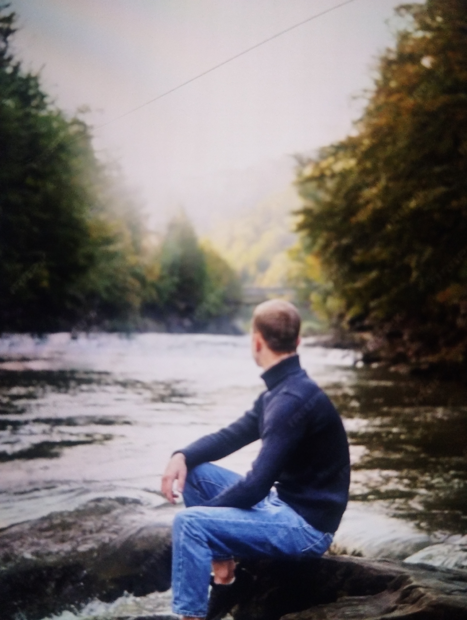 A man is sitting on a rock next to a river, with trees on either side. The man is wearing a dark blue turtleneck sweater and blue jeans. He is looking out at the river. The river is wide and has a moderate current. The trees are a mix of green and yellow, indicating it is autumn. The sky is overcast. This scene is not identifiable as being in Shymkent, Kazakhstan.
