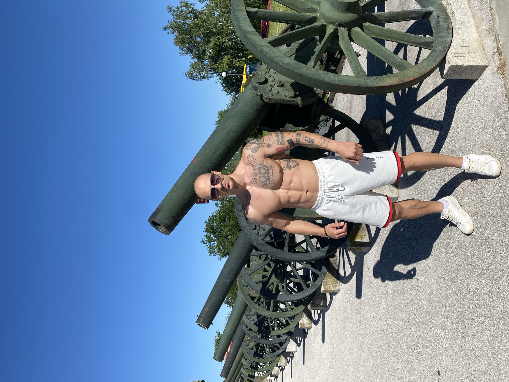 A bald, shirtless man with tattoos on his arms and chest stands in front of a row of cannons in Pleven, Bulgaria. The cannons are green and have large wooden wheels. The man is wearing sunglasses and white shorts with a red stripe on the bottom. He is standing on a concrete surface, and the sky is blue.
