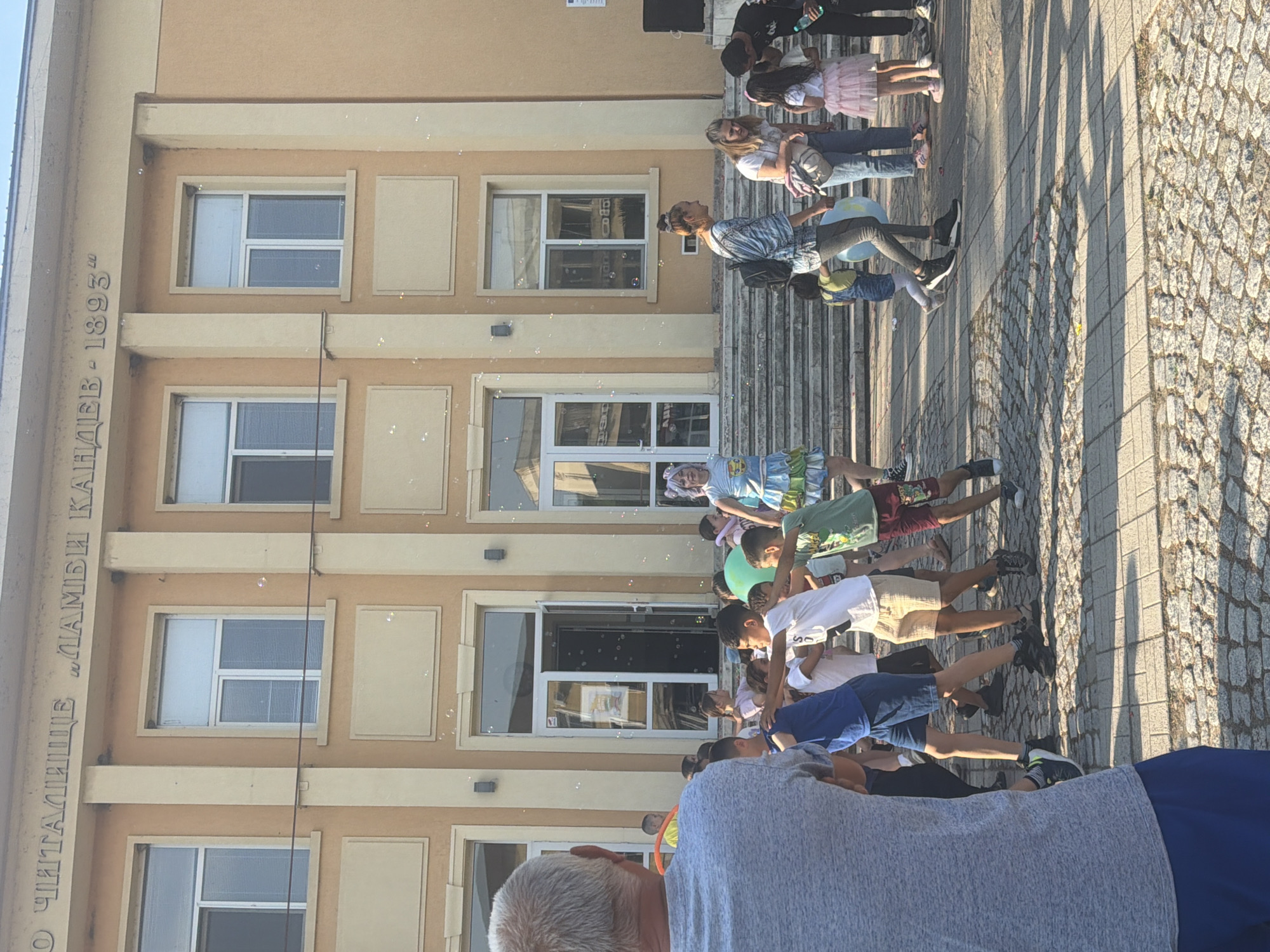 A photo taken in an unknown city in Unknown depicts a gathering of people in front of a building, with numerous soap bubbles floating in the air. The building is three stories tall and has several windows, some of which reflect the surroundings. Inscribed vertically on the building's facade are letters and numbers that read, "ЧИТАЛИЩЕ "ЛАМБИ КАПИДЕВ - 1893"", indicating the building may be a cultural center or library founded in 1893. A set of stairs leads up to the building's entrance, where people stand and observe. A row of individuals, primarily children, are situated on the ground, looking up and seemingly interacting with the bubbles. The foreground shows the back of a person with gray hair wearing a gray shirt and blue shorts. The pavement and the shadows suggest a sunny day with distinct light patterns.