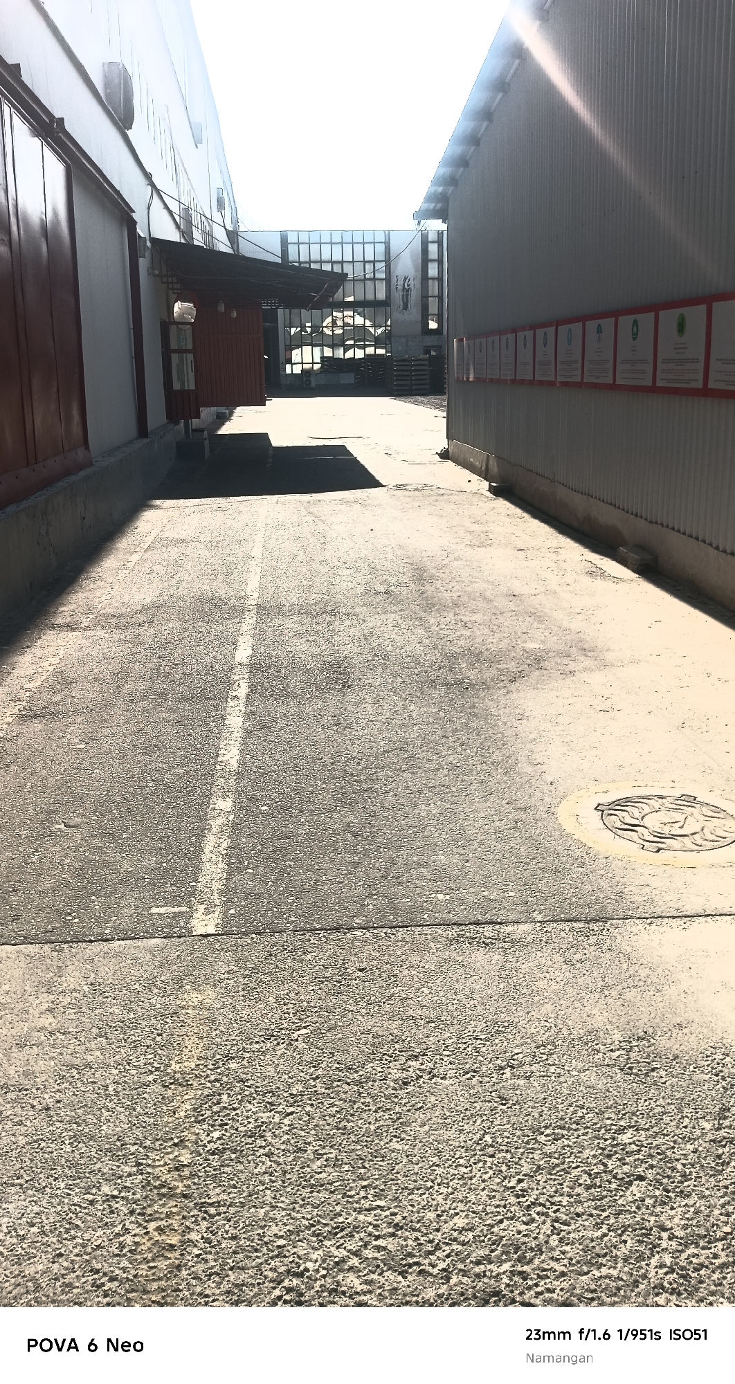 The image depicts an outdoor scene in the city of Namangan, Uzbekistan, likely a service or industrial area. In the foreground, a cracked asphalt ground is visible, marked with a white dashed line. To the left, a building with large, dark red sliding doors is present, with a concrete curb running along its base. To the right, a corrugated metal building runs parallel, with a row of white signs featuring red borders mounted on its exterior. The perspective leads towards a building in the distance with a large glass facade, revealing an interior with industrial elements like wooden pallets. Sunlight casts strong shadows, indicating it is daytime. There are no people or visible animals in the image. The overall impression is one of an unadorned utilitarian space.