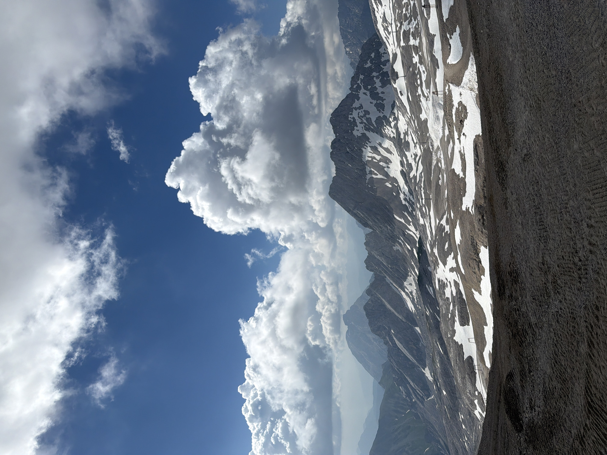 The image shows a panoramic view of a mountainous landscape under a bright blue sky with scattered white clouds. The foreground is a dark, rough terrain, possibly gravel or scree, with visible tire tracks or disturbances. In the mid-ground, jagged mountain peaks rise, sparsely covered with patches of snow and brown earth. Several ski lifts or similar structures are visible on the slopes. The background features more distant, hazy mountain ranges. The overall impression is of a high-altitude, possibly alpine, environment. This landscape is consistent with the Zugspitze massif, the highest peak in Germany, located near Garmisch-Partenkirchen.