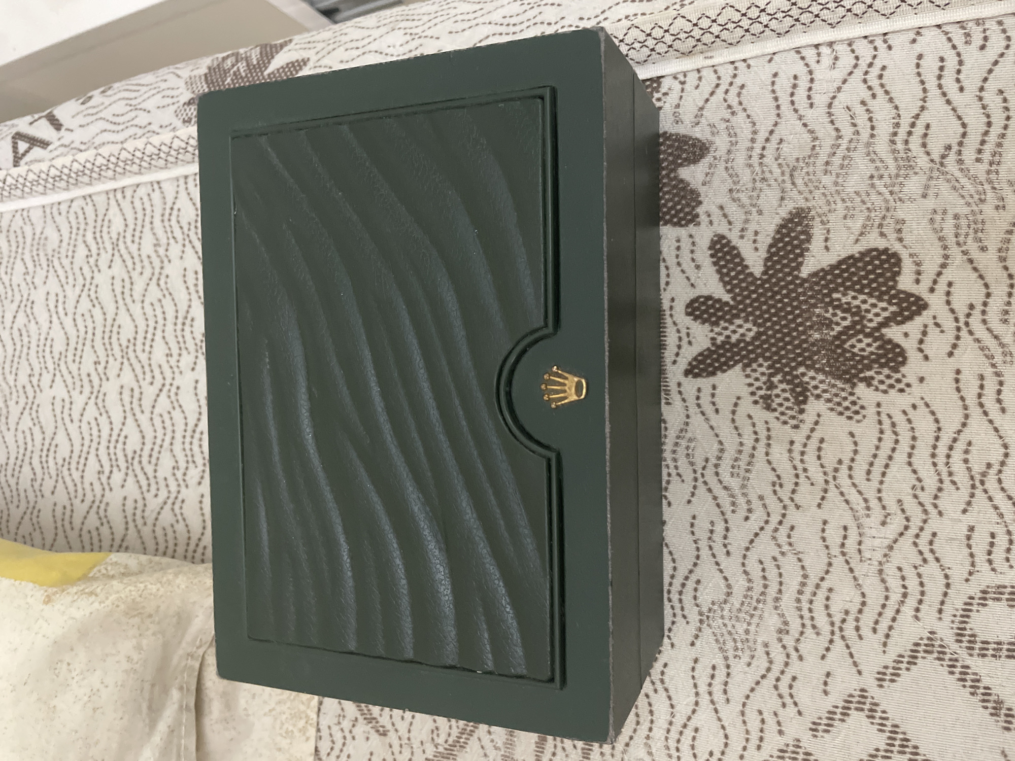 The image displays a dark green rectangular box, identified as a Rolex watch box by the gold crown logo prominently featured on its textured lid. The lid of the box exhibits a distinct wavy, ridged pattern. Slight signs of wear are visible along the box's edges, notably on the top right corner.
The box is placed on a light-colored fabric surface adorned with a repeating pattern. This pattern consists of numerous delicate, wavy brown lines and larger, more solid brown shapes that resemble flowers or stars. In the lower-left corner, a section of another light-colored fabric, possibly yellow or cream, can be seen, suggesting it might be part of a cushion or bedding.
The scene is set indoors, with no human subjects or specific activities depicted. The lighting indicates an interior environment, and neither the time of day nor the weather can be determined from the photograph. While Ihtiman, Bulgaria, is provided as a location context, there are no visual cues within the image itself to corroborate this information. The only visible text is the stylized gold Rolex crown logo on the box.