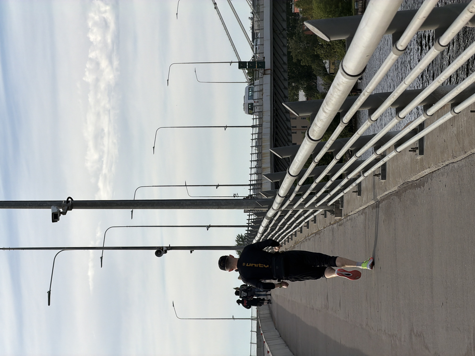 The image depicts an outdoor scene on a wide concrete bridge or elevated walkway, likely in an urban setting. In the foreground, a person is seen from behind, appearing to run or walk. This individual wears a black long-sleeved top with the word "UNITY" emblazoned in yellow text on the back, black shorts, and two different-colored running shoes: one with a red sole and the other with a neon green/yellow upper.
To their left, a series of tall grey poles equipped with streetlights and what appear to be surveillance cameras extend upwards towards a bright, light blue sky with scattered white clouds. To the right, a sturdy metal railing borders the walkway, beyond which a body of water with visible ripples can be seen. Across the water, distant buildings and greenery are visible.
Further along the bridge, behind the main subject, several other people are present, suggesting a group activity like a run or walk. In the mid-ground, on a parallel, higher structure, a white tram or light rail vehicle is discernible. The time of day appears to be bright daylight, indicating clear weather. The scene suggests a dynamic urban environment, consistent with a city like Saint Petersburg, Russia.