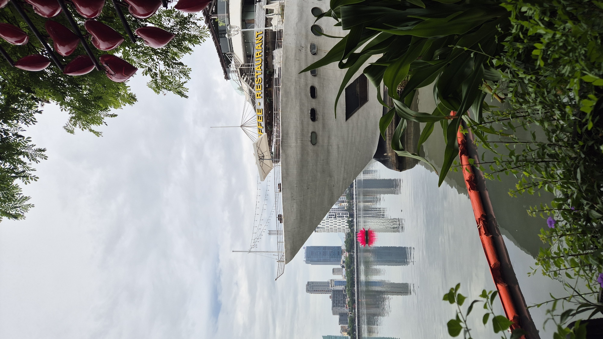 The image captures a daytime scene in Đà Nẵng, Vietnam, likely along the Han River. Dominating the foreground is a large, white, ship-like structure, possibly a restaurant or café, with the text "COFFEE - RESTAURANT" visible in yellow lettering. The vessel has multiple decks, windows, and an umbrella on its upper level. In the distance, a city skyline with several tall buildings is reflected in the calm water. A bright pink, lotus-shaped floating object is positioned in the river between the ship and the city. Green foliage, including large leaves and smaller plants, frames the lower right and bottom edges of the image. The sky is overcast with soft, diffused light. The scene is peaceful and urban, with a focus on the juxtaposition of the man-made structures against the natural elements and the cityscape.