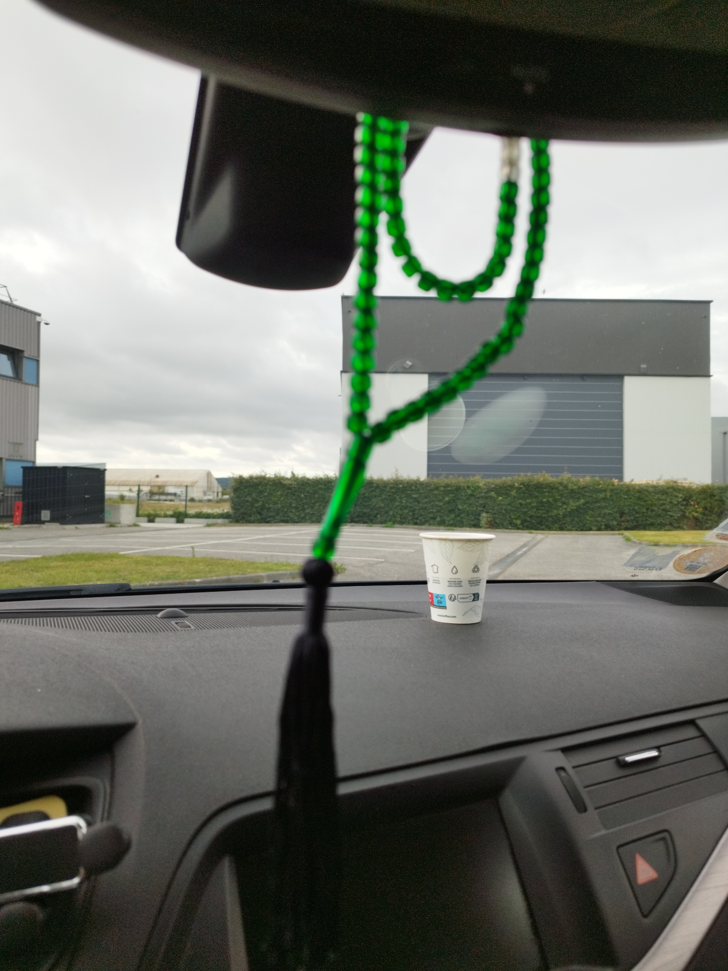 The image is taken from inside a car, looking out through the windshield on a cloudy day. A green beaded necklace with a black tassel hangs from the rearview mirror, partially obscuring the view of the exterior. On the dashboard of the car, a disposable coffee cup is placed. Outside the car, there is a parking lot, a well-maintained hedge, and modern industrial buildings. The image is likely taken in Cherbourg-en-Cotentin, France, given the industrial architecture that is typical of such areas. There are no people visible in the image. The scene is static and depicts a parked vehicle.