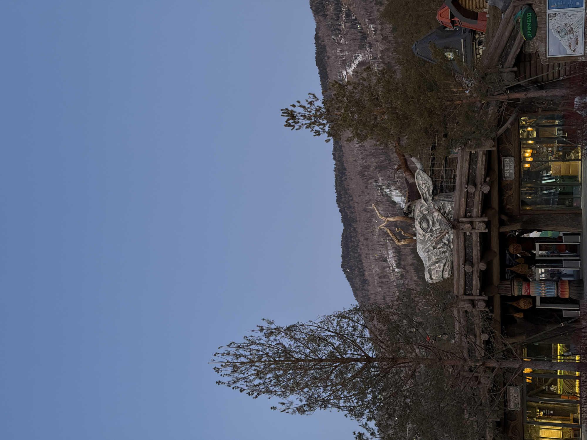 A photograph of a building in Озёрное, Russia, shows a wooden structure decorated with a large, stone moose head. The building appears to be a ticket office with illuminated windows showcasing souvenirs. A forested mountain slopes up behind the structure. Trees partially obscure the view of the building and the mountain. The sky is blue.