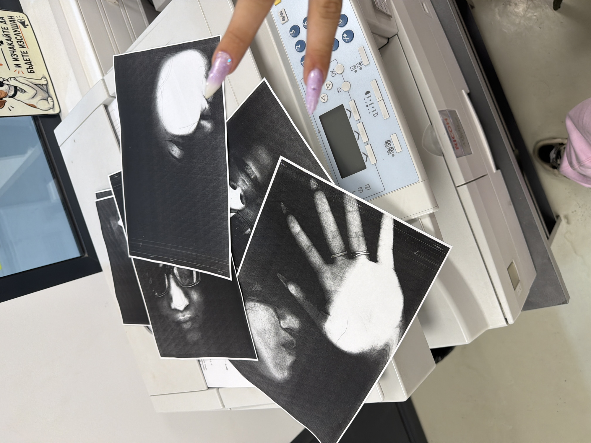 The image displays a close-up, overhead view of several black and white photographs scattered across the top of a white Ricoh copier. The copier is situated on a surface, possibly a desk or table, within what appears to be an office or similar indoor environment. A person's hands with long, light purple, decorated fingernails are visible interacting with the prints and the copier's control panel. One hand is positioned over the copier's buttons, while the other is partially obscuring one of the photographs. The photographs themselves appear to be abstract or artistic, with one prominently showing a hand with rings and another featuring a close-up of facial features. A small sign with an illustration of a dog and text in a foreign language is attached to the top of the copier, to the right of the prints. The floor is visible in the lower portion of the image, with a person's pink pants and a sneaker also present. The scene does not contain any specific visual cues that would allow for a precise determination of its location within Stara Zagora, Bulgaria.