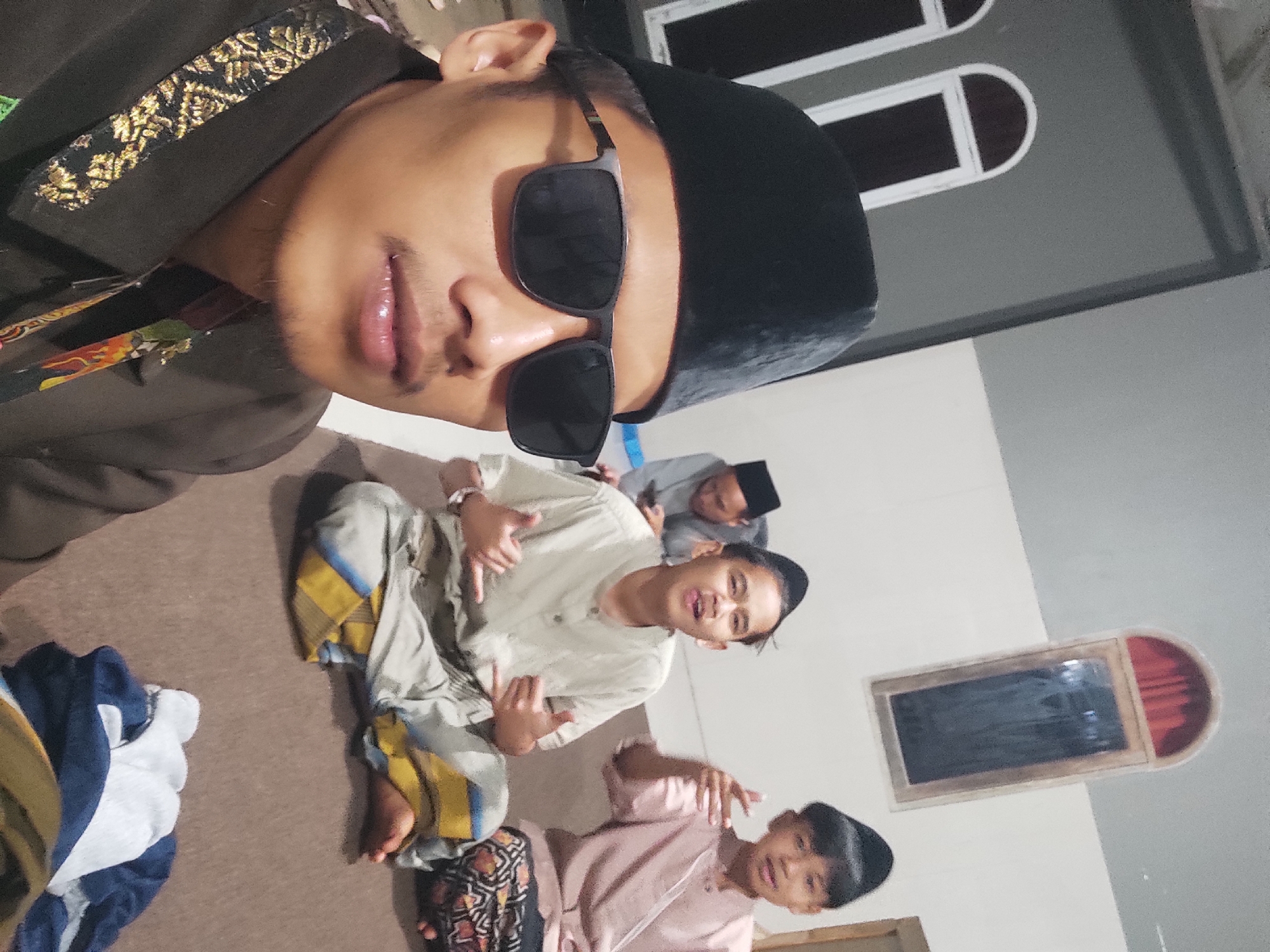 The image captures a group of four young males wearing traditional Islamic attire, including peci hats. The foreground is dominated by a close-up of one individual wearing sunglasses and a dark brown jacket with ornate gold trim. Behind him, three other males are seated on a brown carpeted floor. One is partially visible in the background, wearing a grey shirt. The two in the mid-ground are more prominent: one in a light-colored, collared shirt and patterned sarong, making a gesture with his hands; the other in a pinkish garment and a patterned sarong, also gesturing. The environment appears to be an indoor space, possibly a mosque or a community hall, with a plain grey wall and arched windows visible in the background. The lighting is dim, suggesting it might be evening or an interior setting. There are no clear indicators of the specific location within Sumedang, Indonesia, beyond the general assumption of an Indonesian context given the attire.