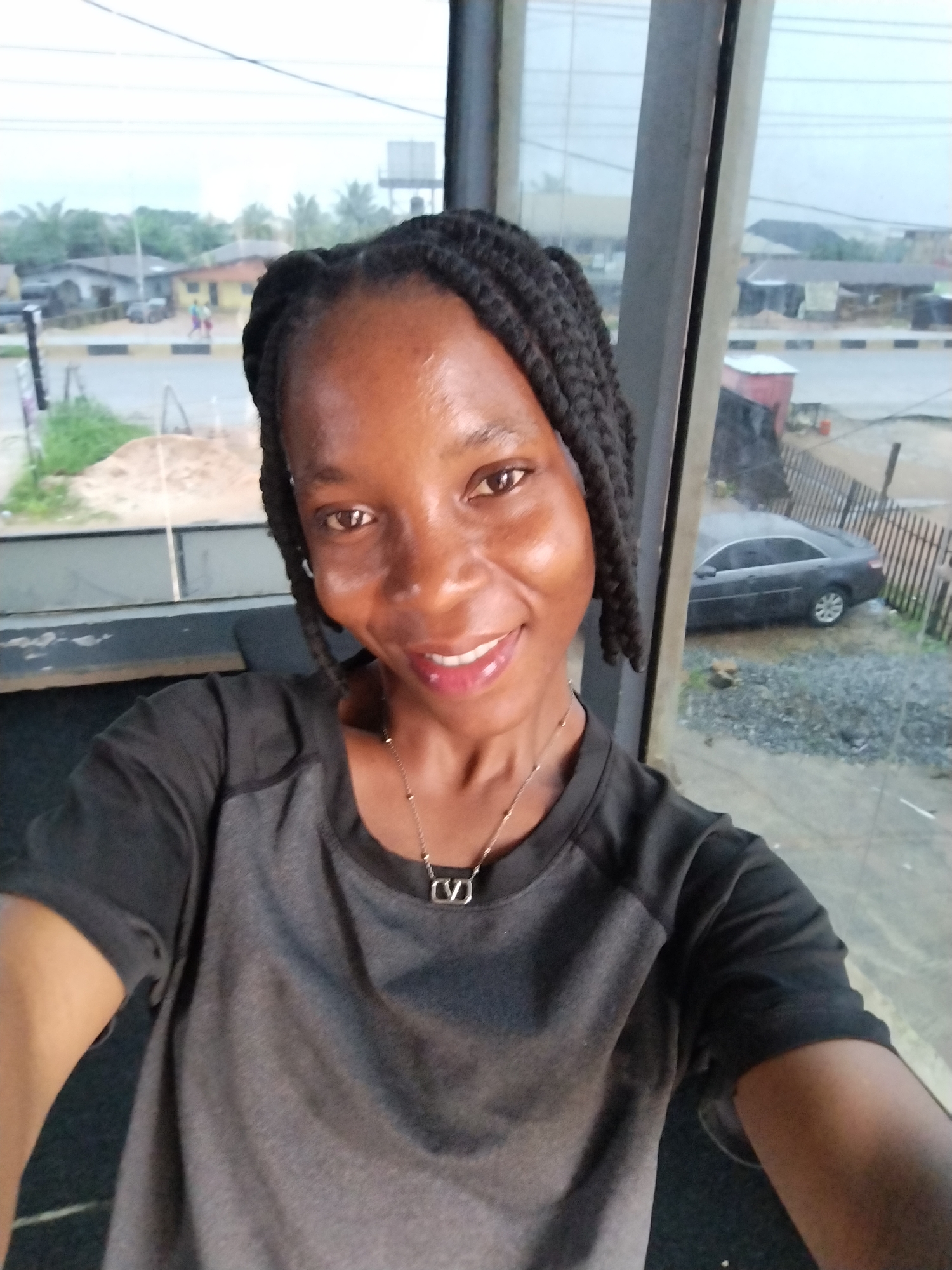 In Uselu, Nigeria, a dark-skinned woman with braided hair is taking a selfie. She is wearing a dark grey t-shirt and a silver necklace with a pendant. Her face is turned towards the camera, and she is smiling. The background shows a street scene with buildings, a car, and trees, possibly indicating an outdoor or semi-outdoor location. The lighting suggests it may be overcast or late in the day.