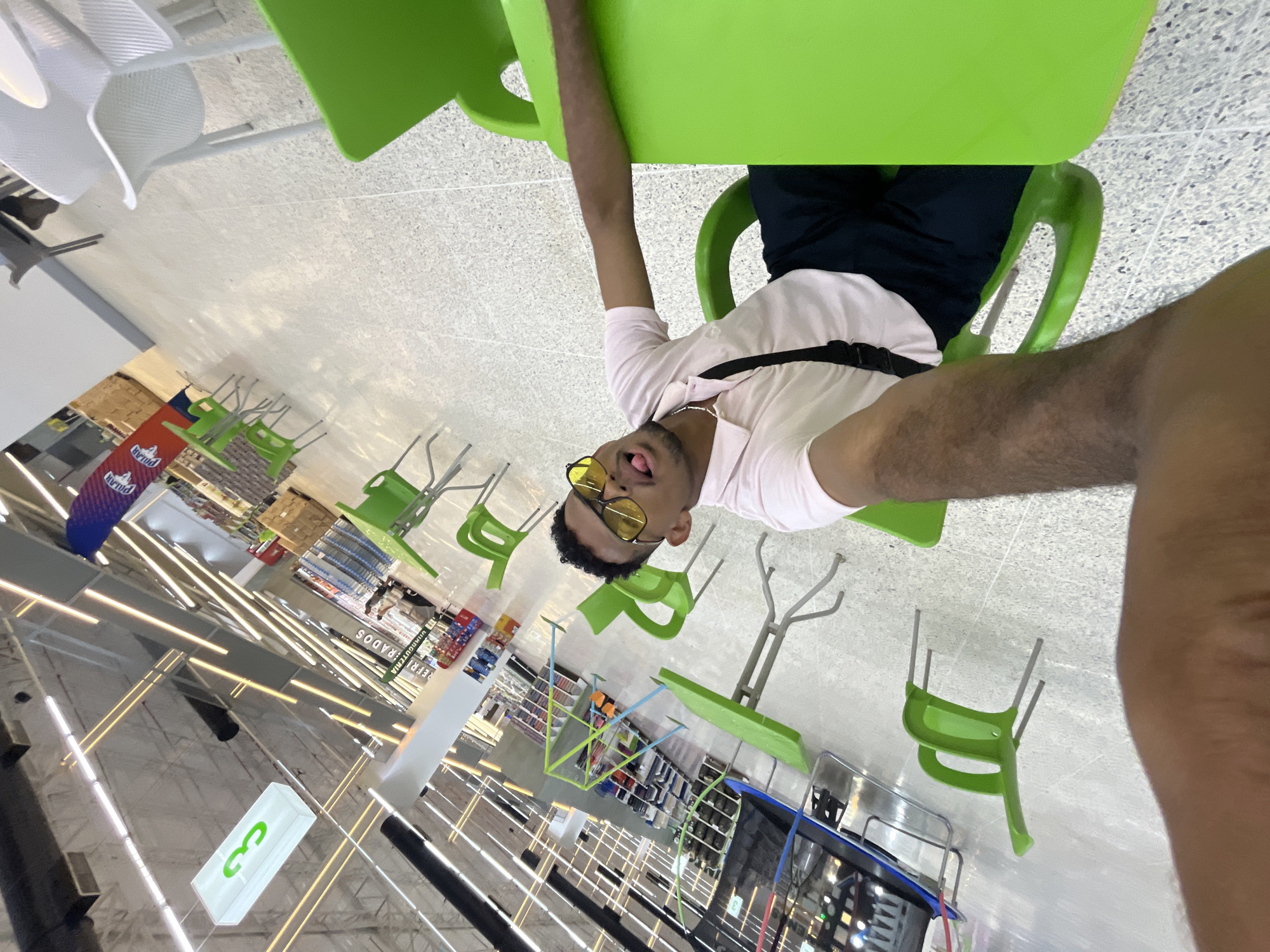 A man wearing a white shirt and yellow sunglasses is sitting at a green table in a supermarket. He is partially visible from the waist up and is sticking his tongue out. The supermarket, identified by logos as belonging to a commercial chain in Venezuela, is well-lit with rows of shelves stocked with products. Other green chairs and tables are seen in the area, suggesting a seating or dining space within the supermarket. The location of the supermarket is suggested to be in Porlamar, Venezuela.